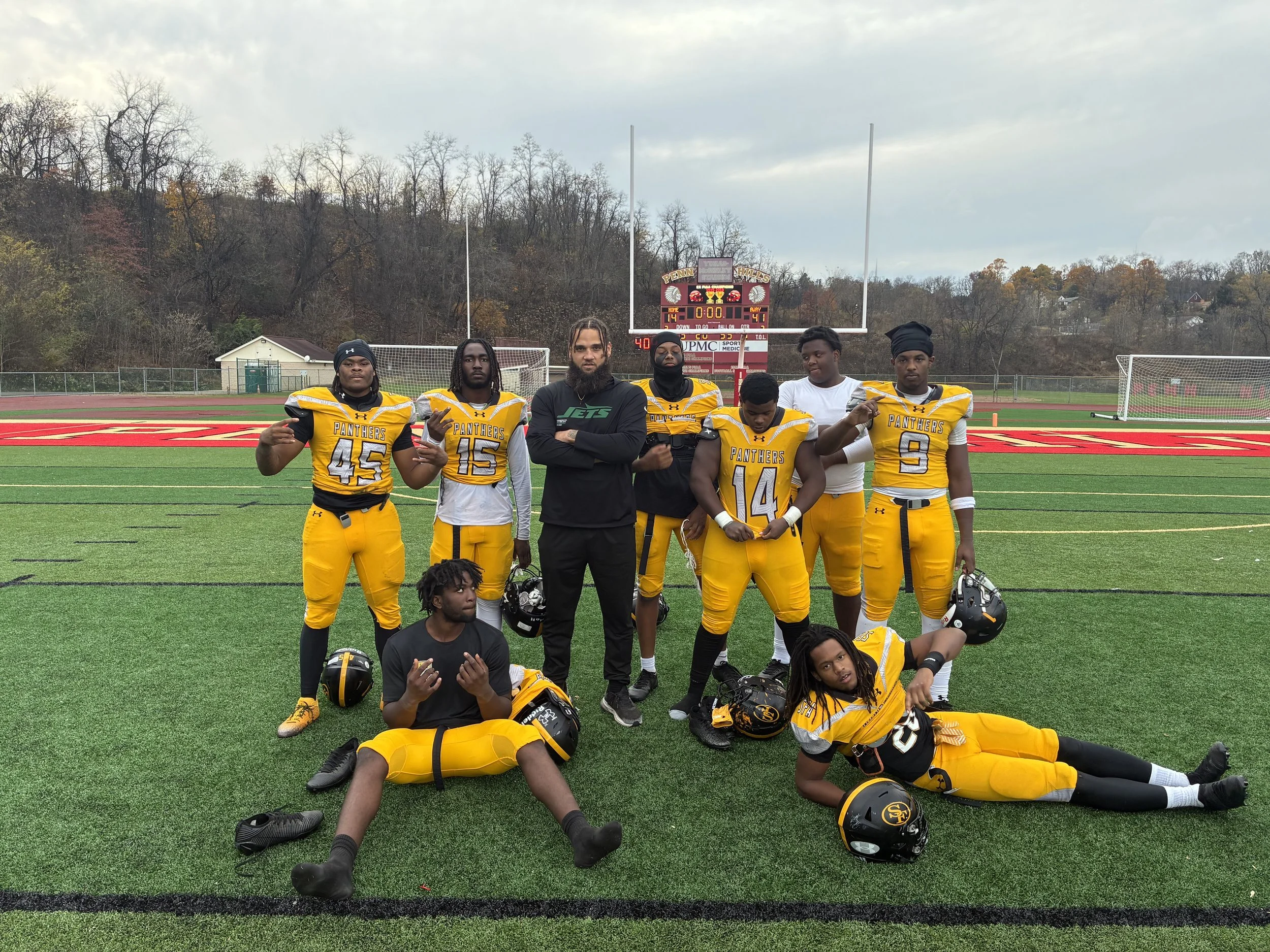 A group of football players standing and sitting on a football field with a scoreboard in the background. They are wearing yellow and black uniforms with 'Panthers' written on them. Some players are holding helmets, and others pose with peace signs or crossed arms.