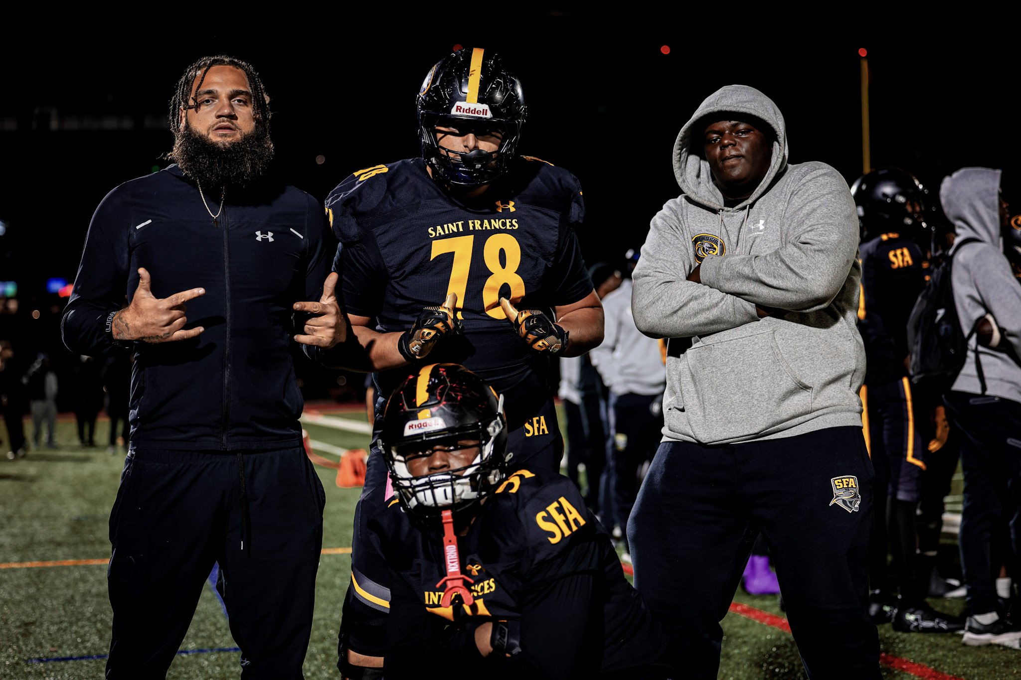 Four football players, two in black and yellow uniforms and two in gray hoodies, stand on a field at night. One player is kneeling with a helmet. The others are standing with arms crossed or pointing at each other.