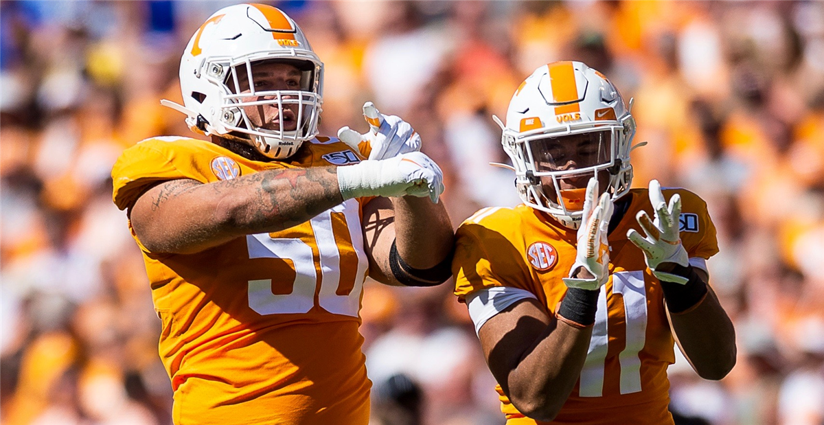 Two American football players in orange and white uniforms celebrating on the field, with a blurred crowd in the background.