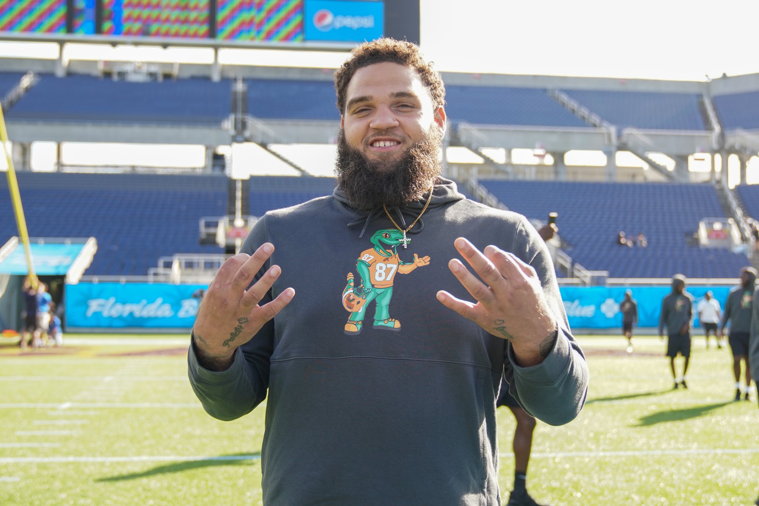 A smiling man with a beard stands on a football field in an empty stadium, wearing a gray hoodie with a cartoon alligator mascot and the number 87. He is making a gesture with both hands, and there are people in the background.