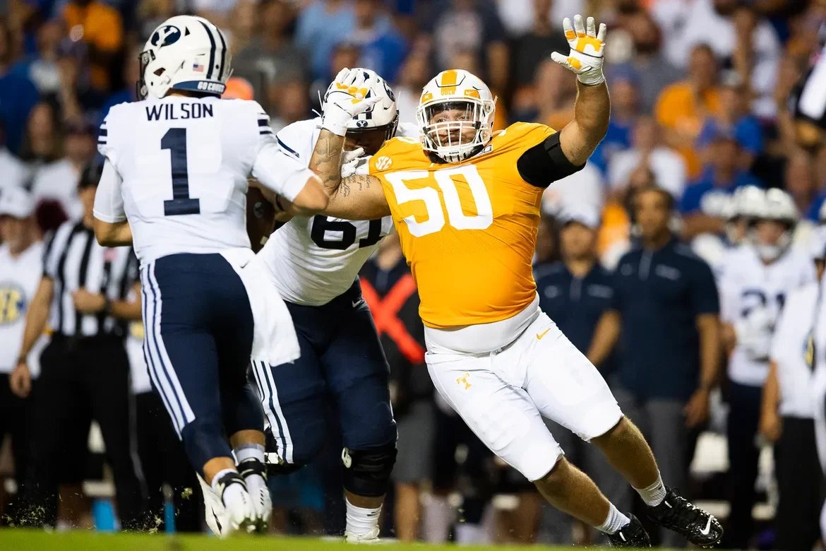 American football players in action during a game, with one player in a yellow jersey number 50 and two players in white jerseys, one with the name Wilson on the back, engaged in a play on the field with spectators in the background.