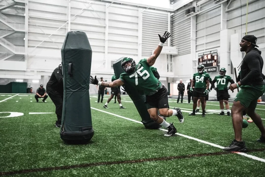 Football players practicing drills in an indoor training facility, with one player wearing a green jersey number 75 blocking a padded dummy.
