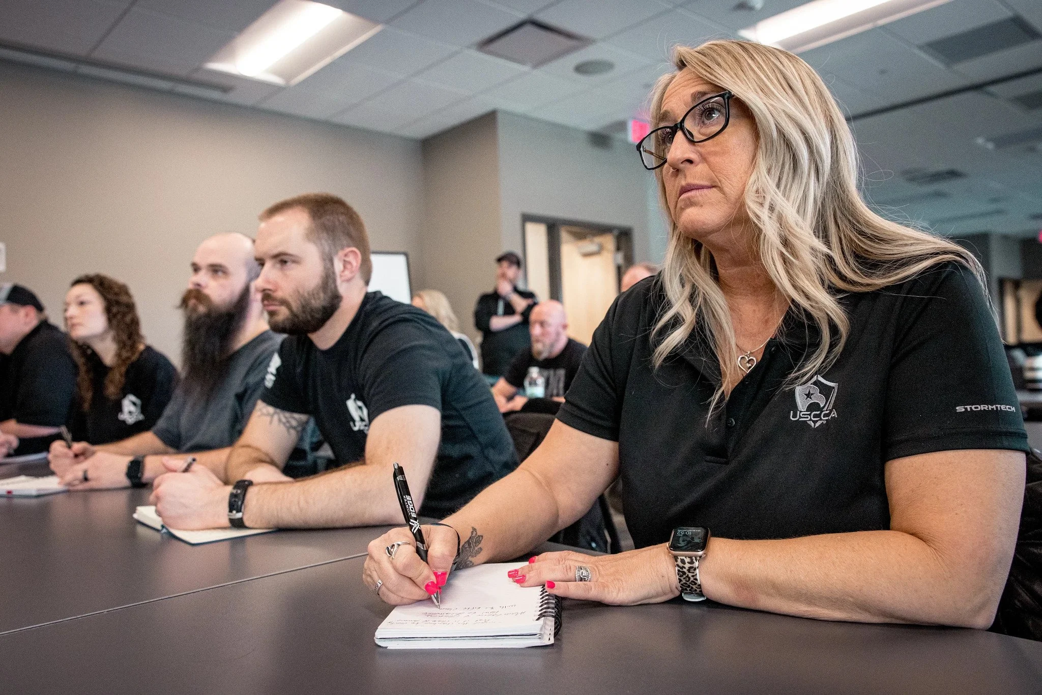 Group of people attending a meeting or seminar, sitting at tables, listening attentively, taking notes, with a woman in the foreground wearing glasses, a black polo shirt with a patch, and writing in a notebook.