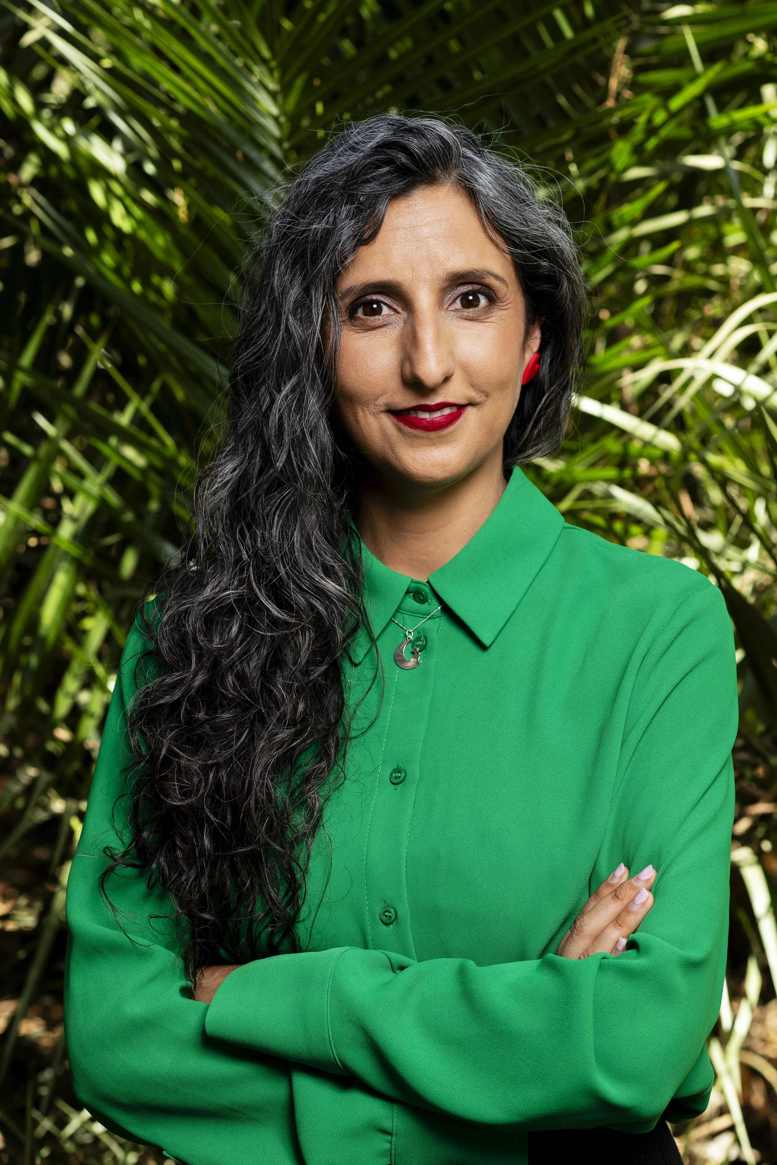 Mujer con cabello rizado y gris, vistiendo una blusa verde, frente a un fondo de plantas verdes, con los brazos cruzados y sonriendo.