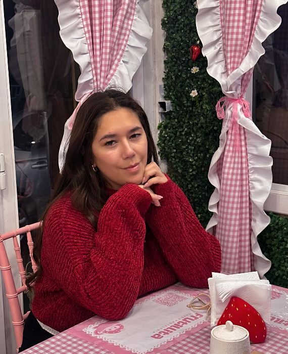 Joven mujer con sudadera roja sentada en una mesa con mantel de cuadros rosas y blancos, en un ambiente decorado con cortinas a cuadros con volantes y fondo de plantas verdes.