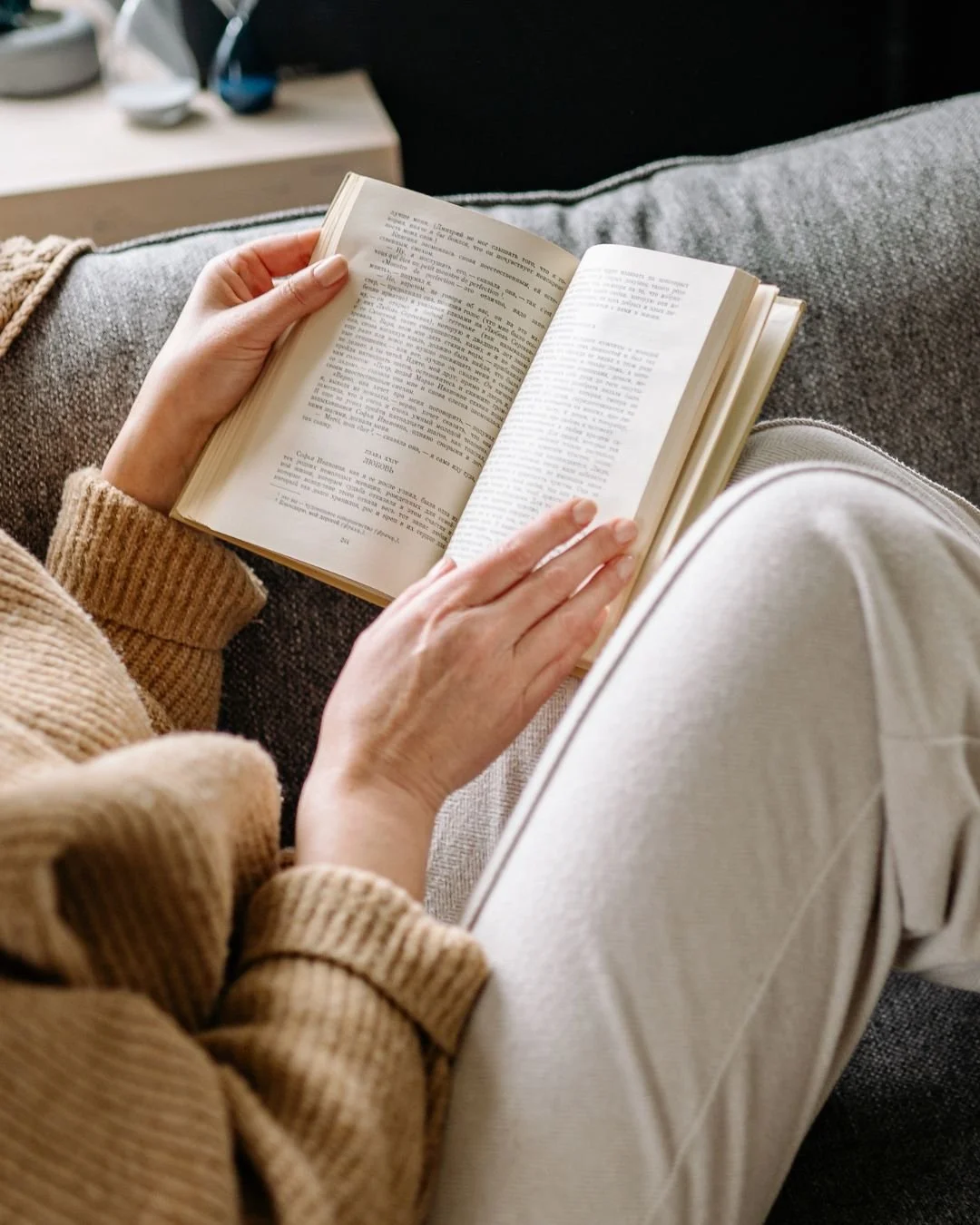 Persona sentada en un sofá leyendo un libro con las piernas cruzadas, llevando un suéter marrón y pantalones beige.