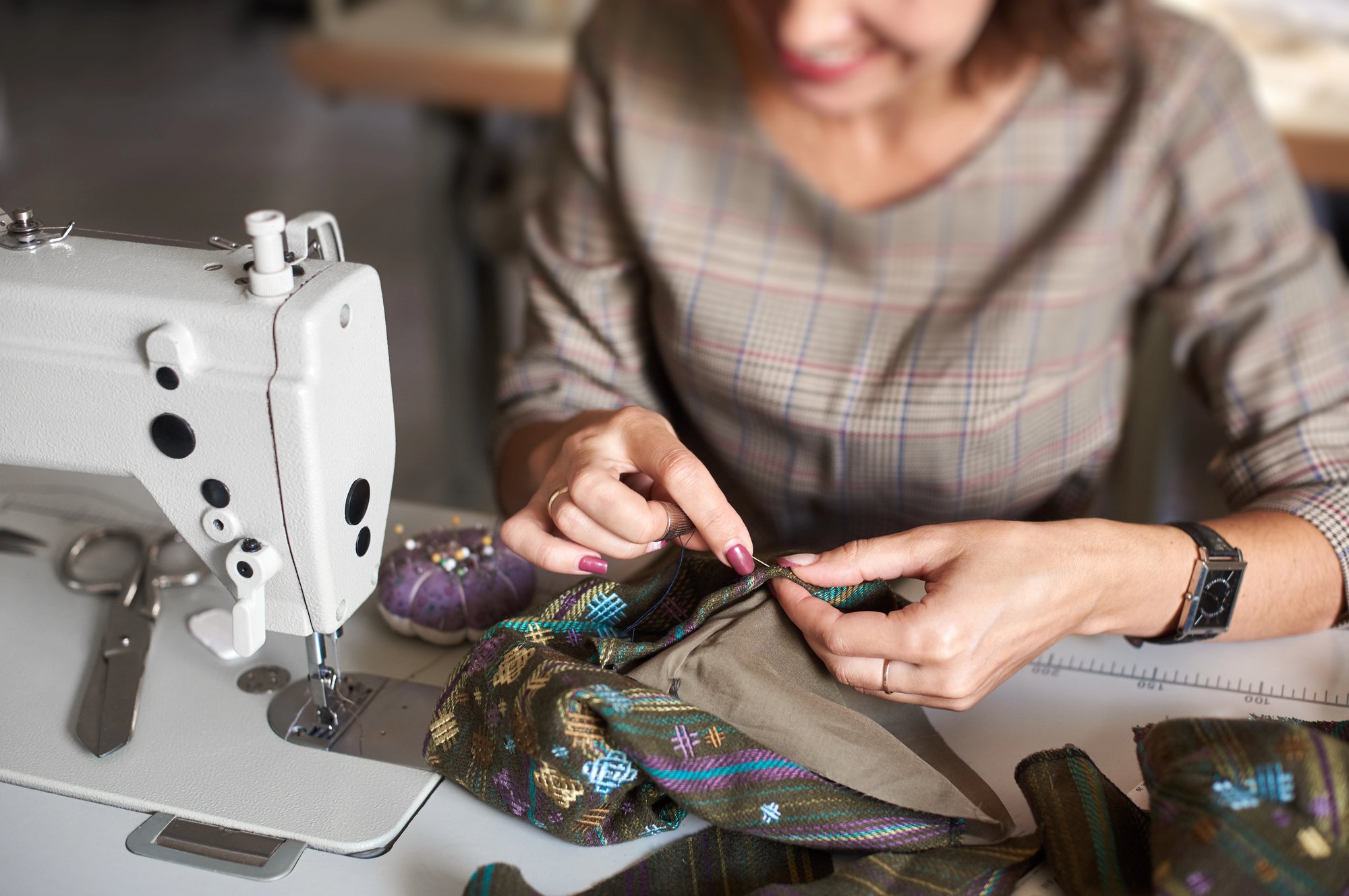 Una mujer sonriente cose con una máquina de coser mientras trabaja con tela de colores y patrones. Está enfocada en su tarea, con las manos en el proceso de costura.