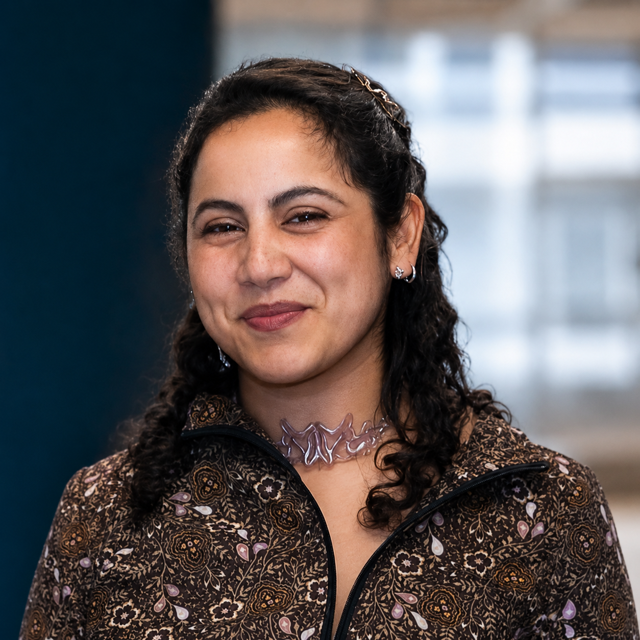 Retrato de una mujer joven con cabello rizado, usando un collar de plástico y pendientes plateados, sonriendo suavemente.