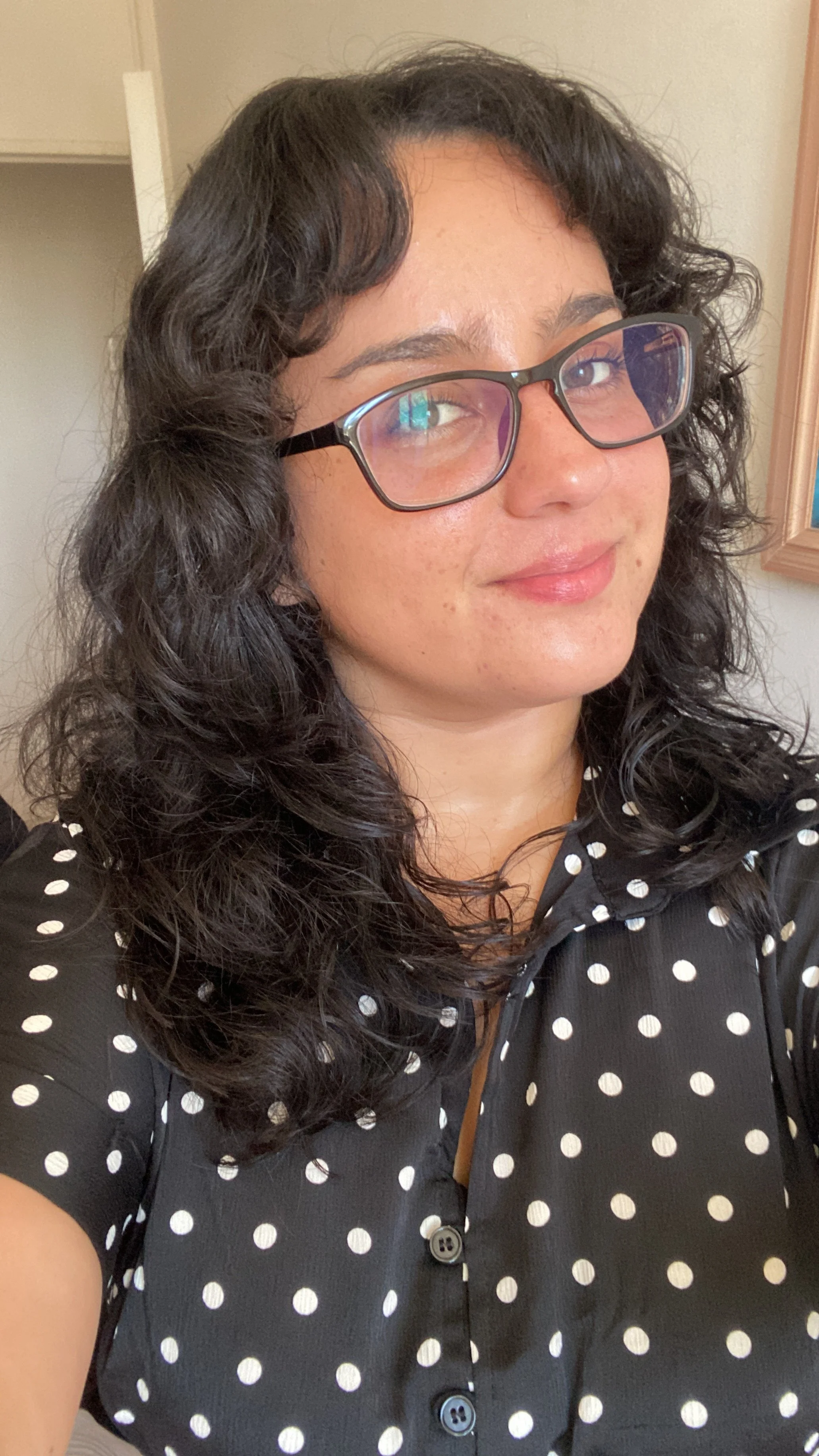 Mujer de cabello rizado negro y lentes con marco negro, vestida con blusa negra de puntos blancos, sonriendo en un espacio interior.