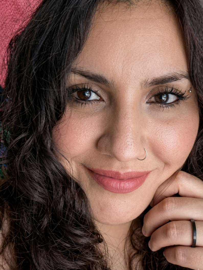 Retrato de una mujer con cabello oscuro rizado, piel clara, luciendo maquillaje natural, con un pendiente en la nariz, con una expresión amistosa, tocándose la barbilla y sonriendo sutilmente.