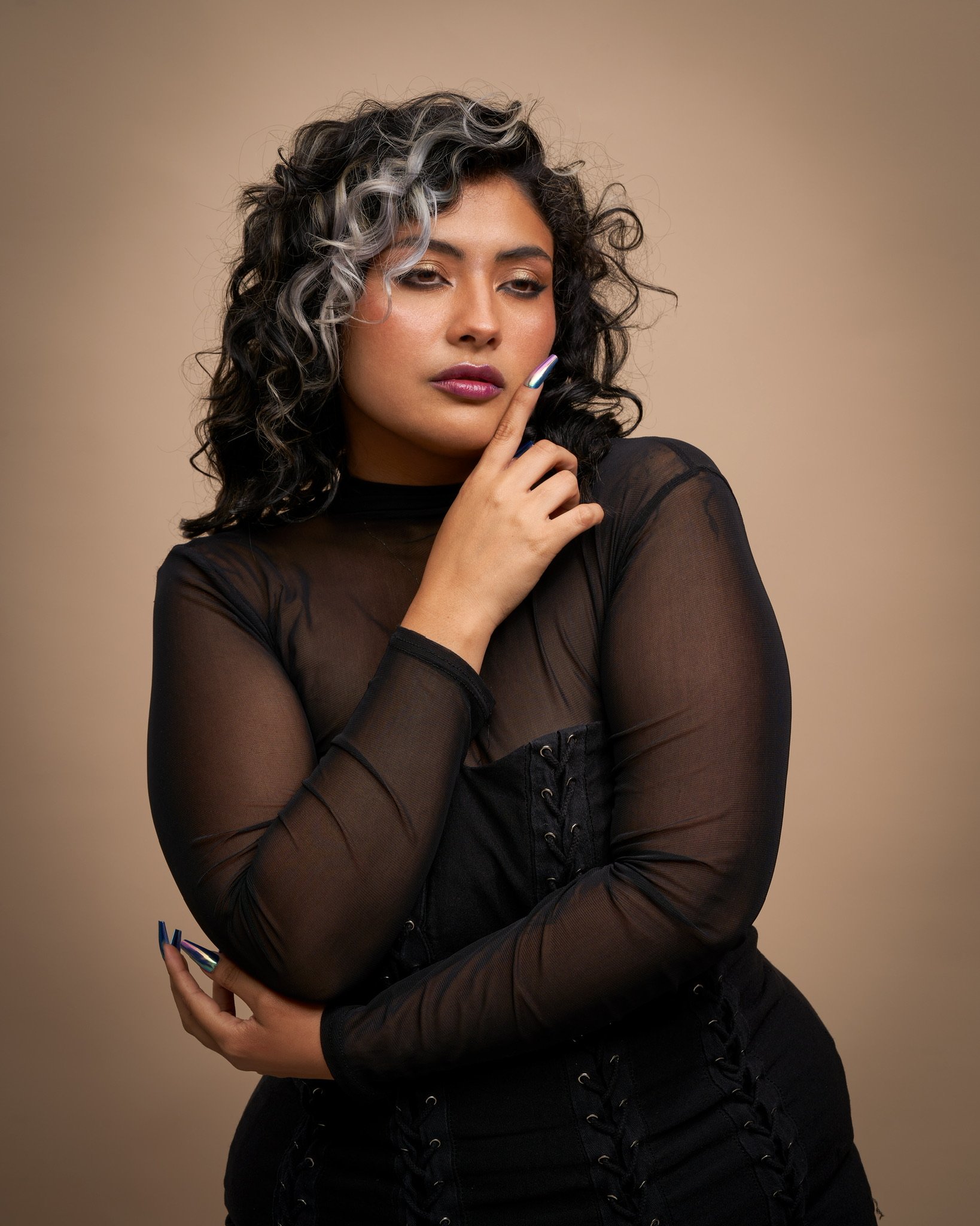 Retrato de una mujer con cabello rizado y canoso en las puntas, vestida con ropa negra de manga larga y transparente, con maquillaje de ojos dorado y labios rosados, posando con expresión pensativa sobre un fondo beige.