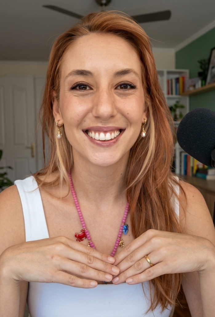 Mujer con cabello rojo, sonriendo frente a la cámara, en un ambiente interior con estanterías y plantas, junto a un micrófono.