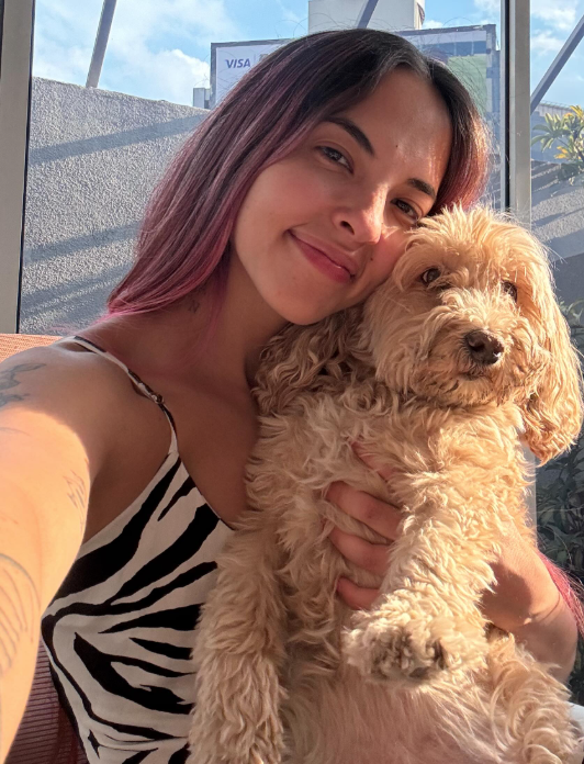 Una mujer con cabello teñido de color rosa y cabello natural, sonriendo y sosteniendo a un perro de raza pequeña, con pelaje rizado y de color crema, en un espacio con ventanas grandes y cielo visible.