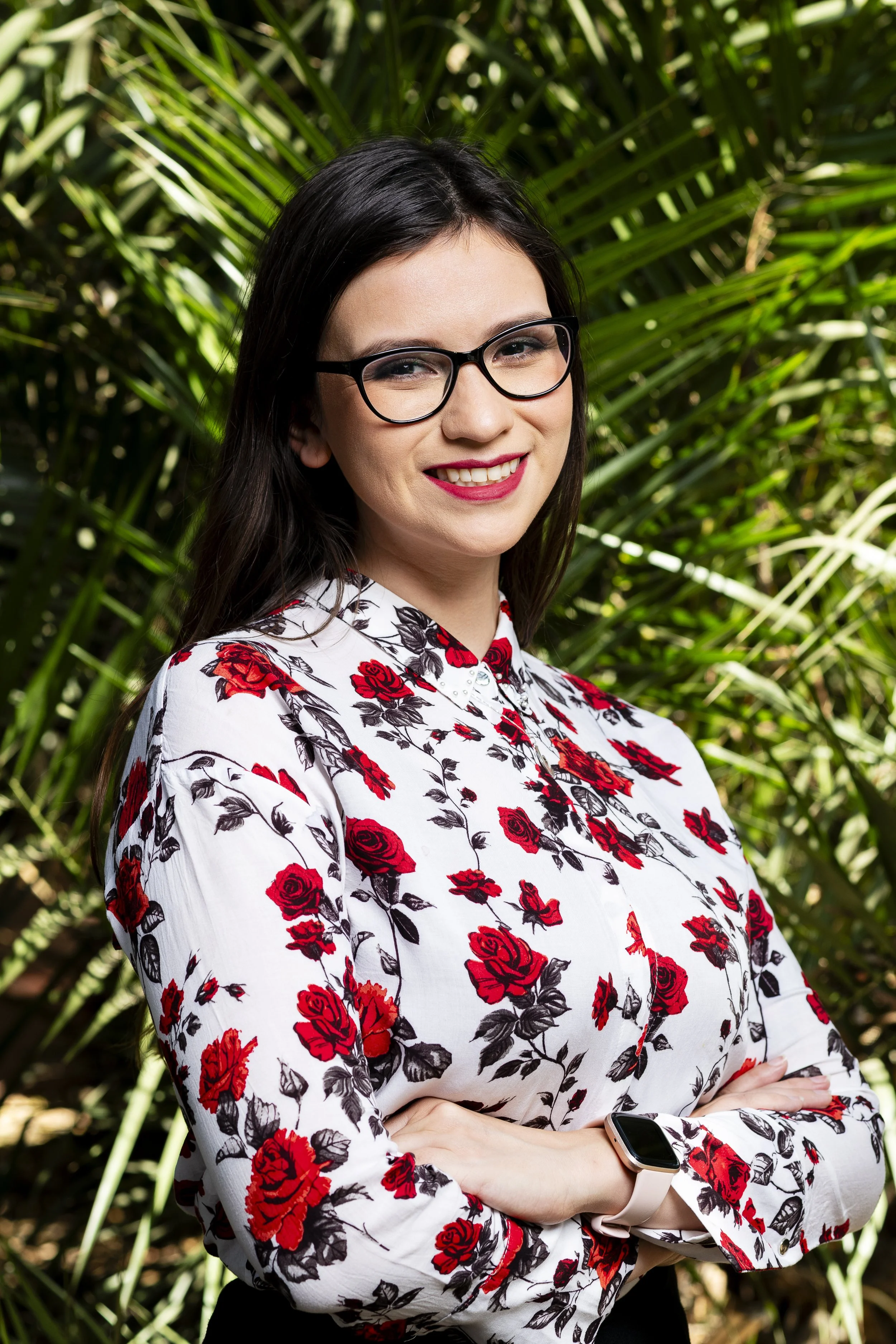 Mujer con gafas y vestido con estampado de rosas rojas y negras, sonriendo y con brazos cruzados, en un ambiente natural con plantas verdes.