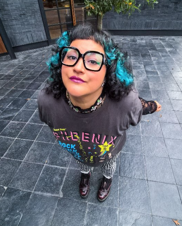 Joven con cabello rizado con puntas teñidas de azul, gafas grandes, camiseta con diseño colorido, pantalones a rayas, y zapatos oscuros, de pie en un piso de baldosas negras, mirando hacia arriba.
