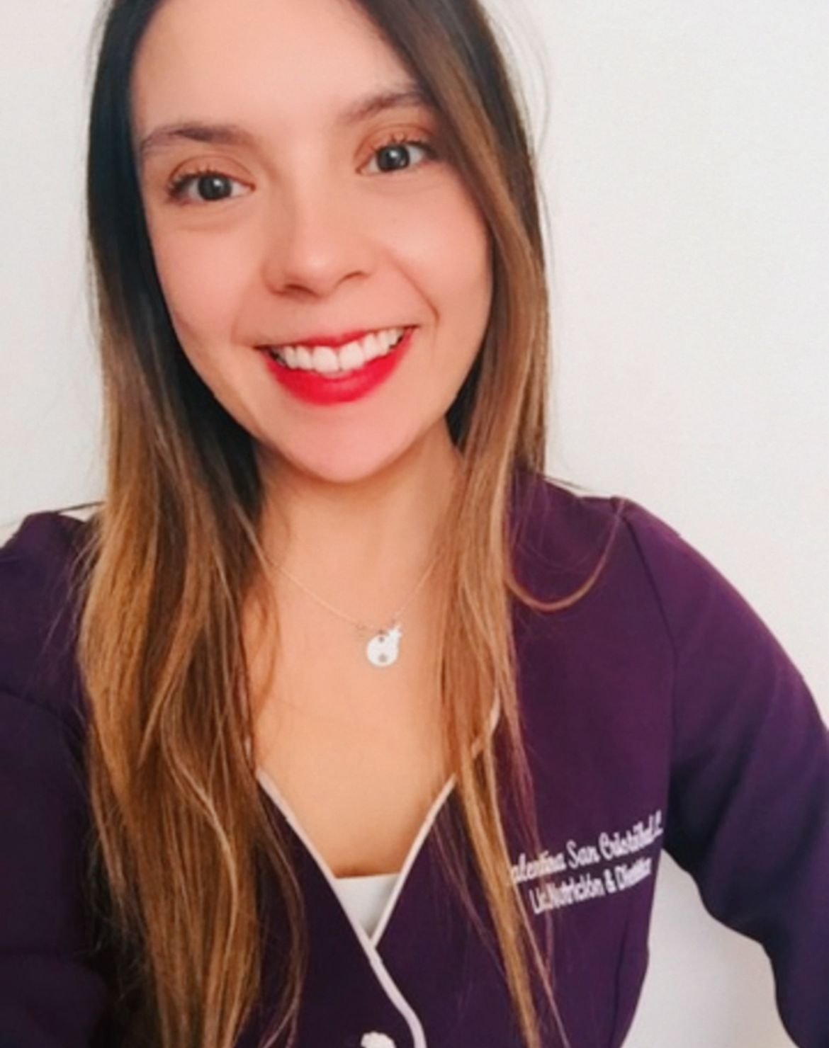Una mujer sonriendo, con cabello largo y rubio, usa uniforme de trabajo de color púrpura y lleva un collar con una medalla de gato.