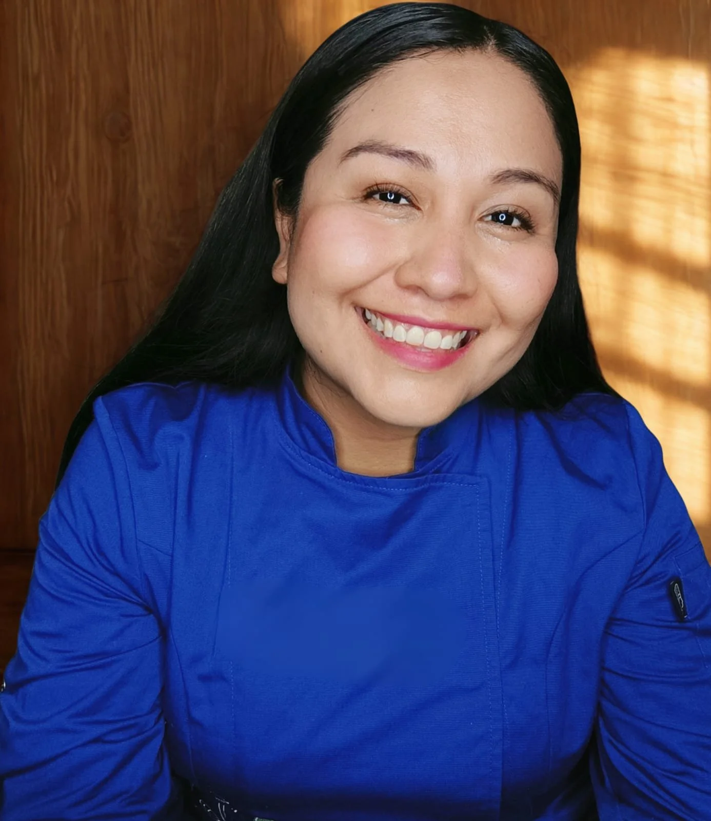 Mujer sonriente con cabello largo y negro, lleva una chaqueta azul y está frente a una pared de madera y cortinas con luz.
