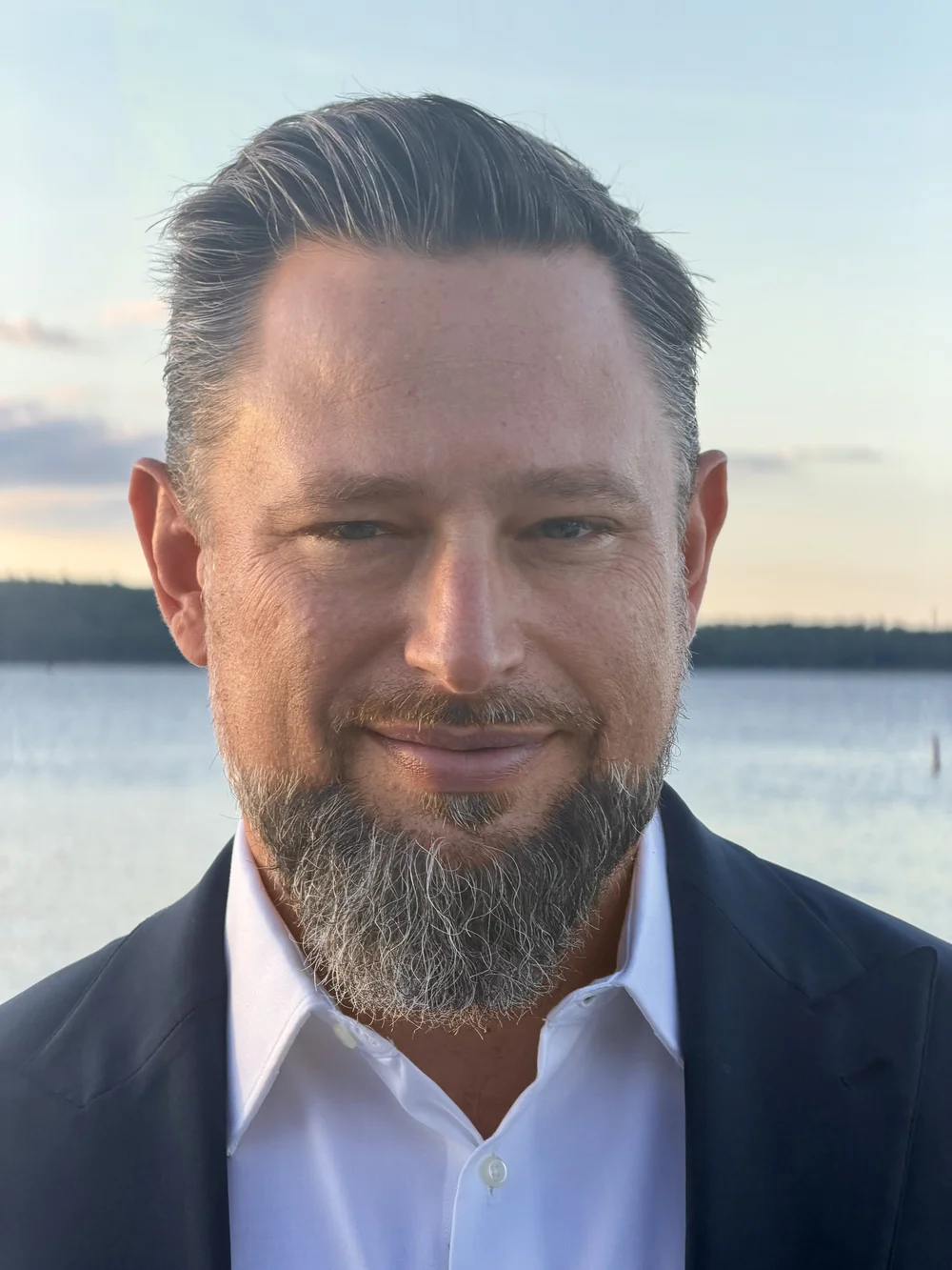 Doug Vanisky in a white shirt and dark blazer, standing outdoors near the water.