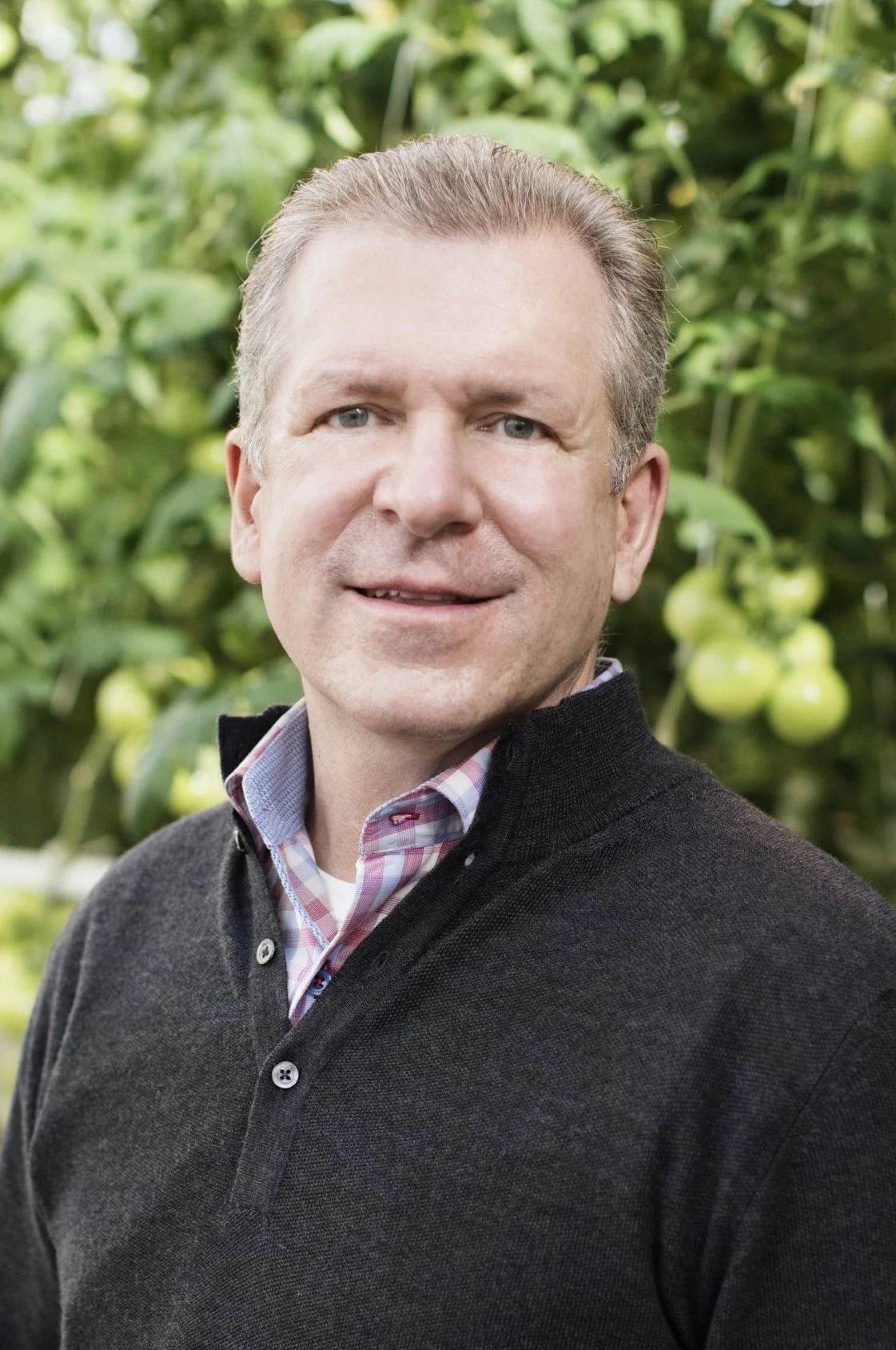A man with short, light brown hair and blue eyes standing outdoors with green foliage and tomato plants in the background.