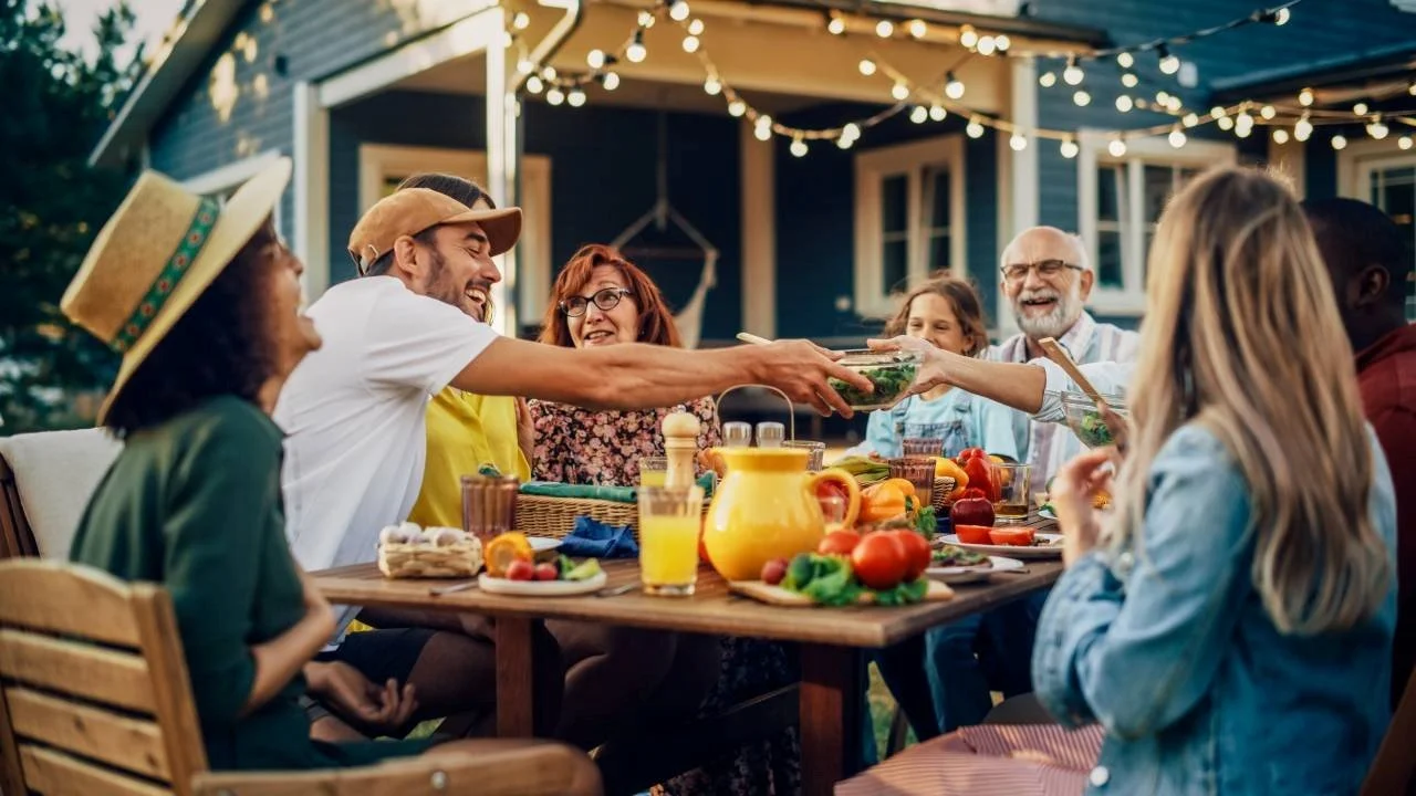 People smiling and sharing food outdoors at a backyard gathering during evening with string lights overhead.