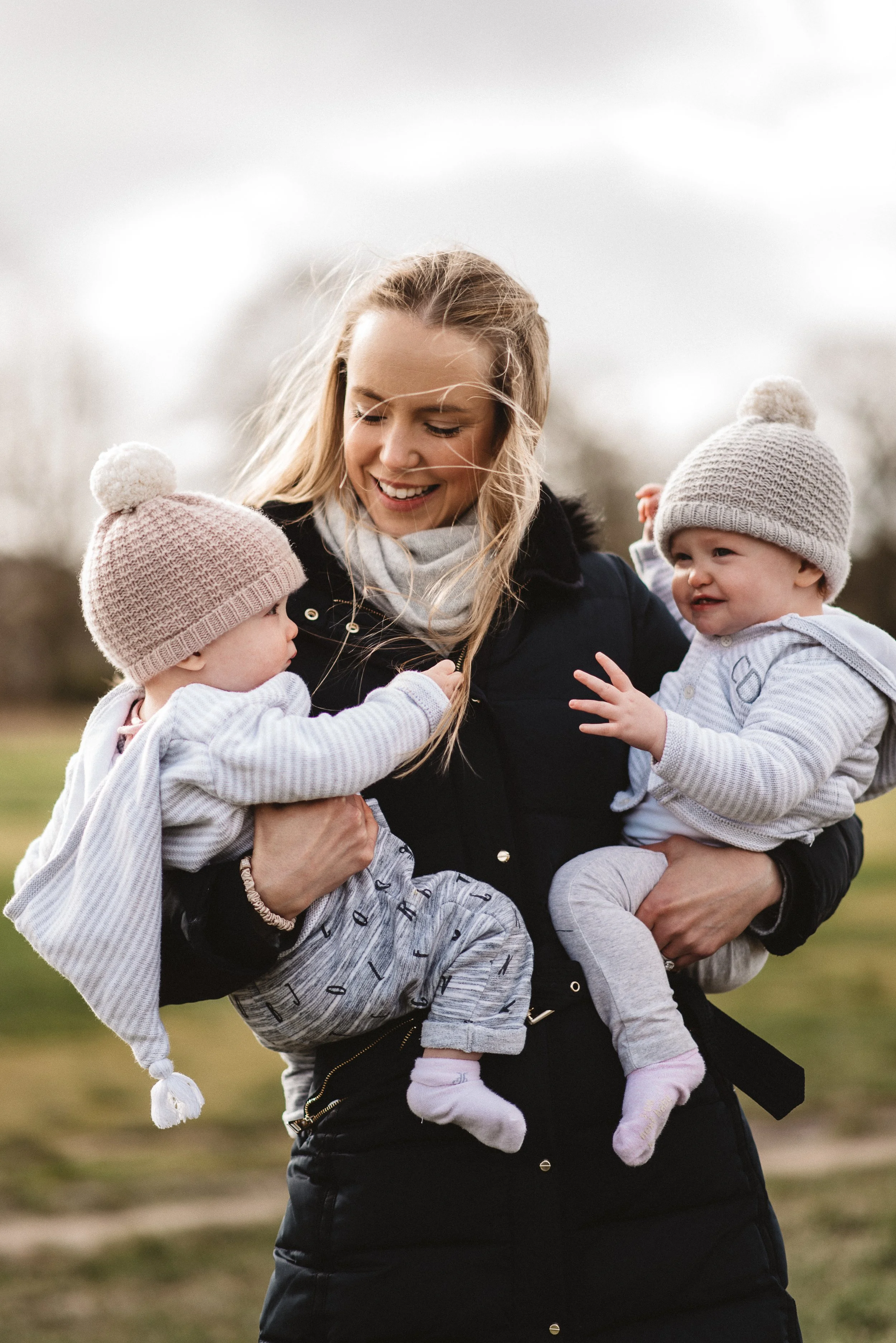 A woman holding two babies, one in each arm, outdoors on a cloudy day. The woman has blonde hair and is smiling, the babies are dressed in knit hats and light-colored clothes.