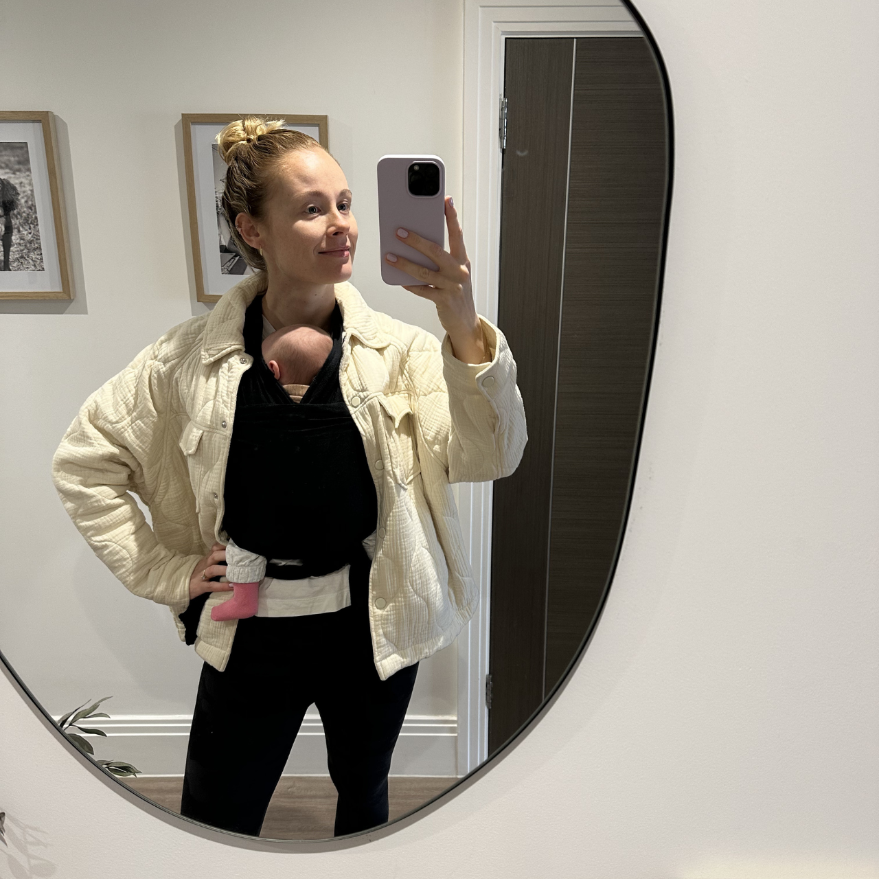 Woman with red hair in a bun taking a mirror selfie, holding a baby in a black carrier on her chest, in a room with framed art on the wall.