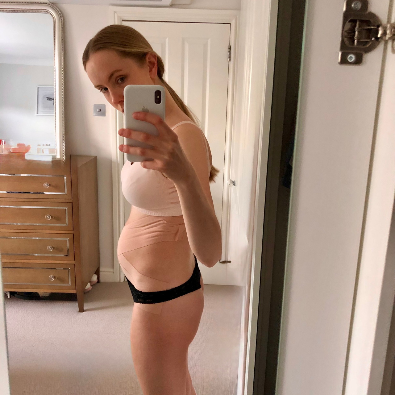 A woman taking a mirror selfie in a bedroom, wearing a beige sleeveless top and black underwear, with her hair tied back.