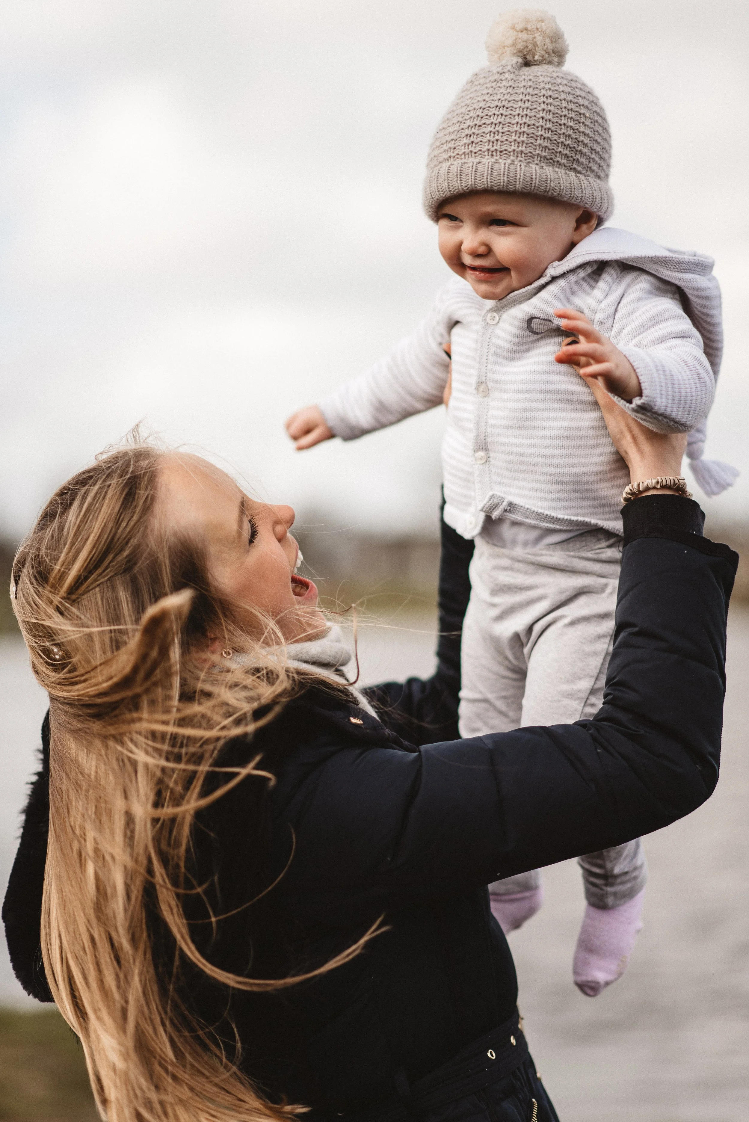 A woman with long blonde hair, wearing a black jacket, lifts a smiling baby dressed in gray and a knit hat high above her under an overcast sky outdoors.