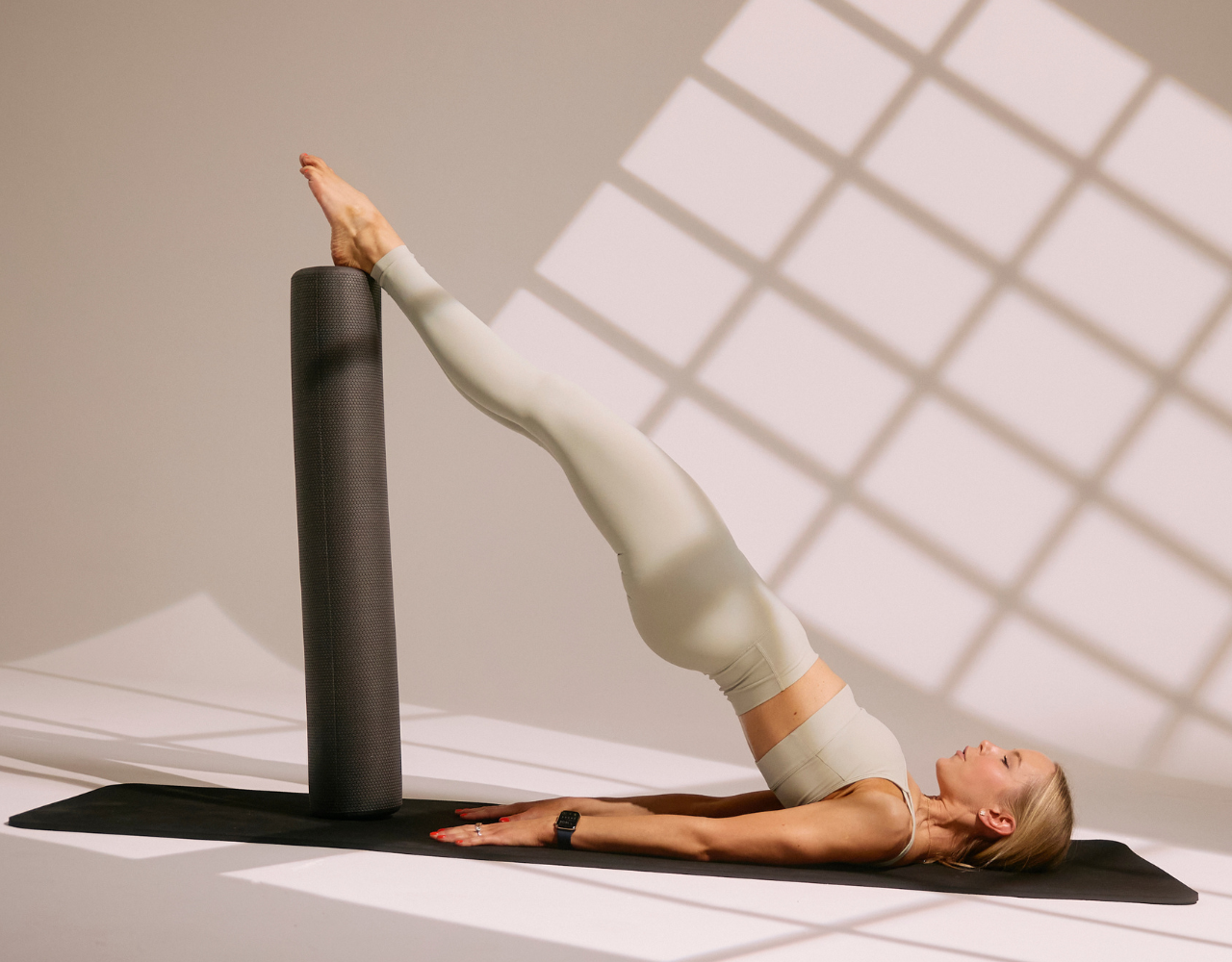 A woman doing yoga on a black mat, with her feet elevated on a foam roller, in a room with sunlight creating shadows on the wall.