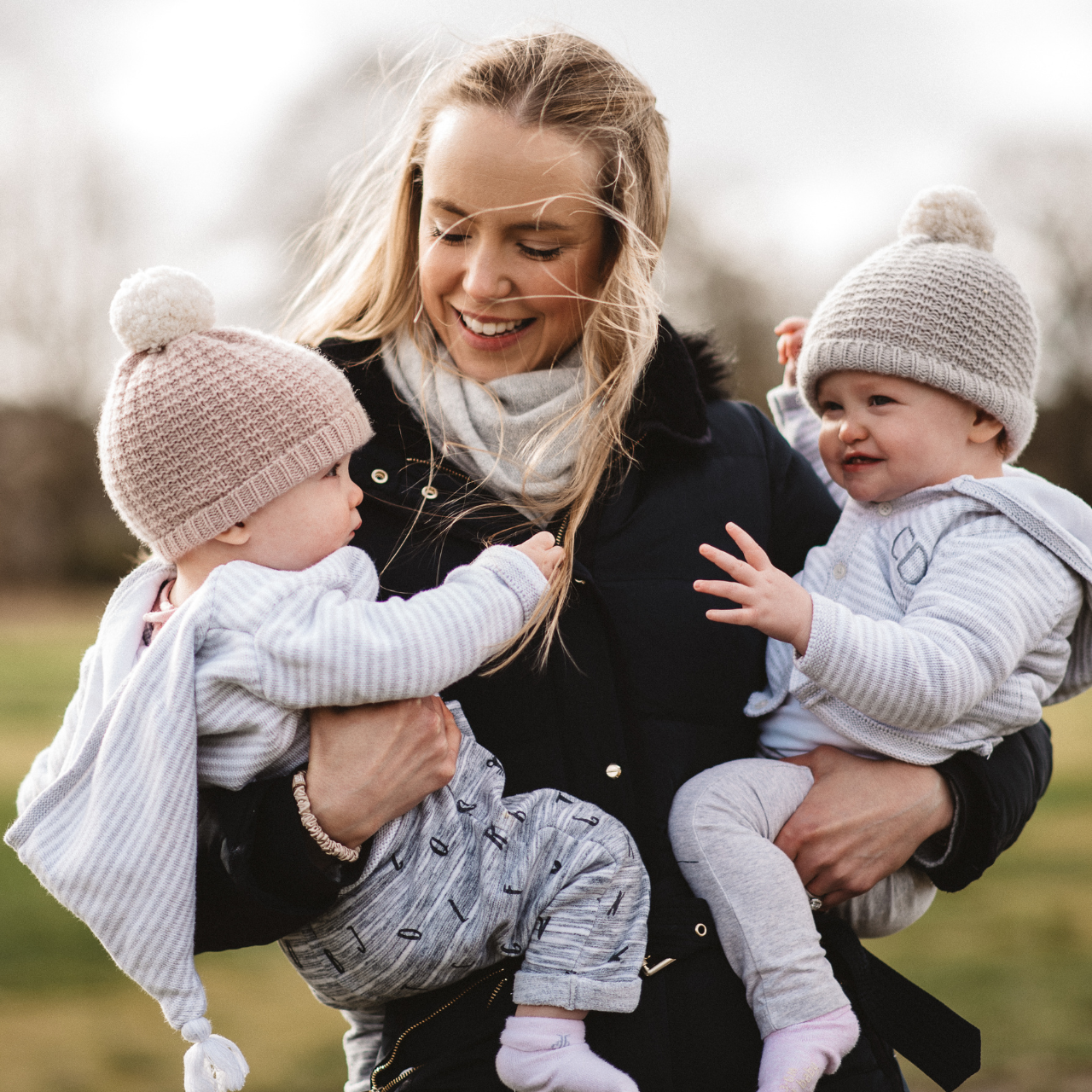 A smiling woman outdoors holding two young children, both wearing knit hats and cozy clothes.