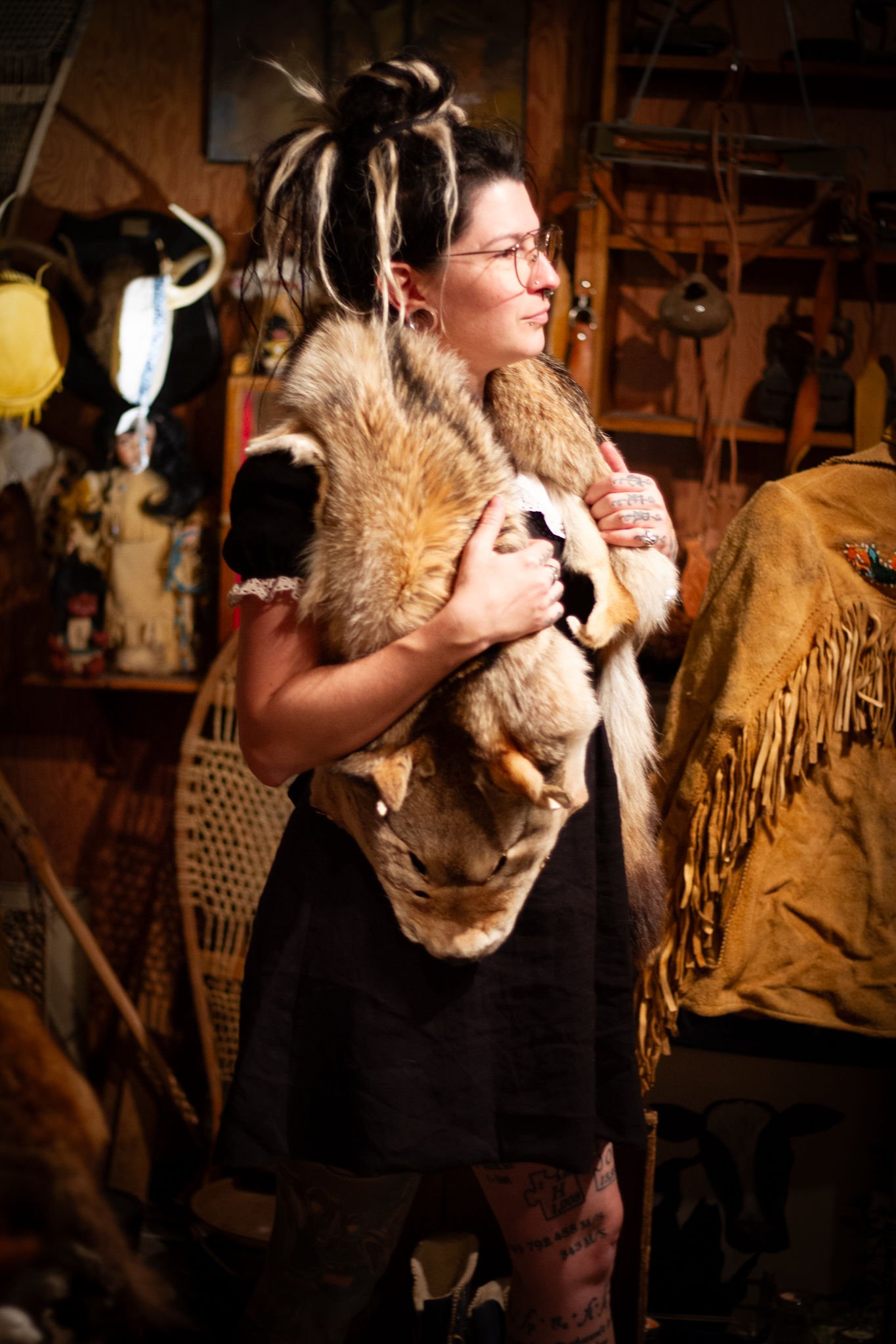 Une femme avec des dreadlocks, porte un manteau en fausse fourrure et des lunettes, dans un intérieur en bois avec des objets artisanaux et des sacs en arrière-plan.