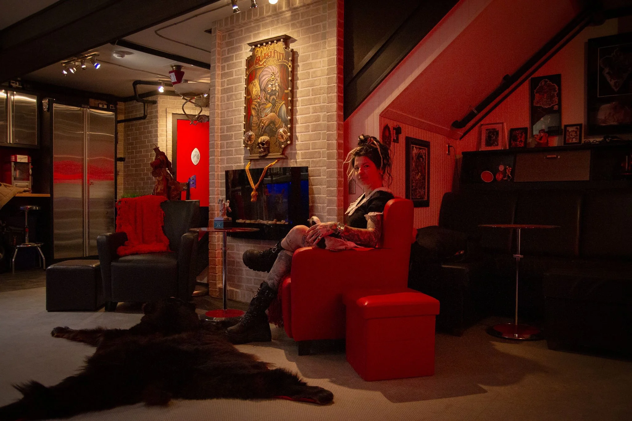 Une femme assise dans un salon, portant un tatouage sur la jambe, avec un meuble de cheminée derrière elle, ambiance tamisée et décorations murales.