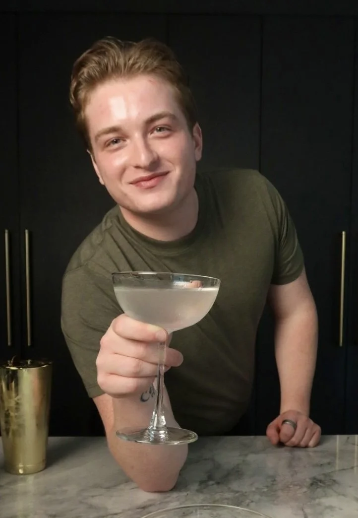 A young man with short light brown hair smiling and holding a cocktail glass filled with a clear drink, standing behind a marble countertop in a dark-colored kitchen.