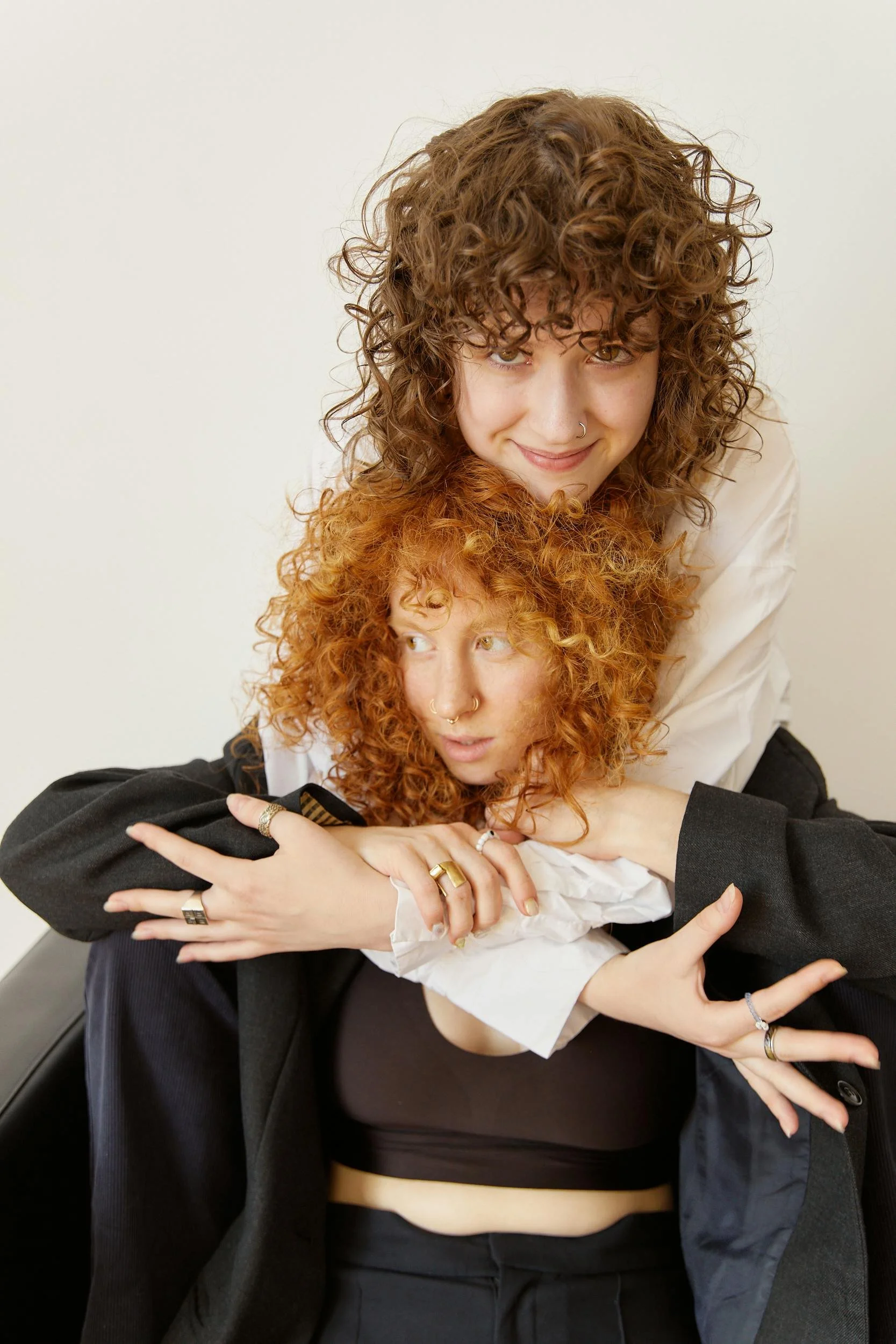 Deux femmes aux cheveux frisés, l'une debout derrière l'autre, avec des expressions complices, portant des vêtements décontractés.
