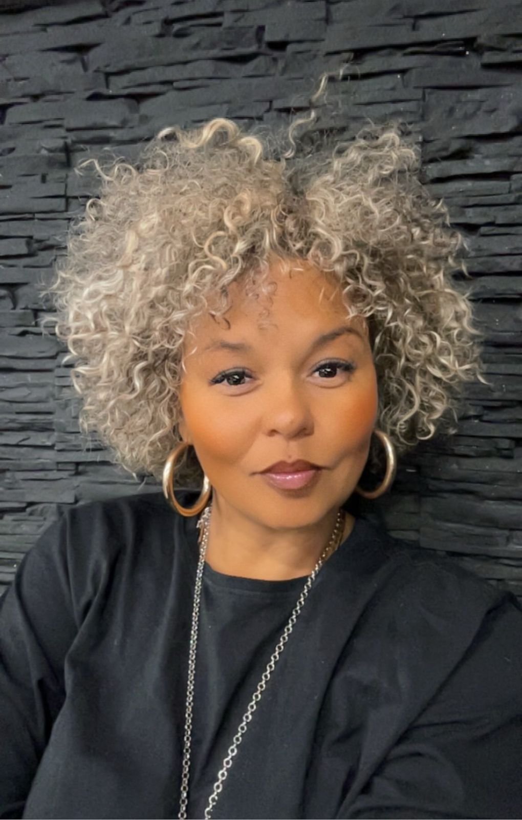 Portrait d'une femme avec des cheveux bouclés gris et blonds, portant des boucles d'oreilles en argent, un collier, vêtue d'un haut noir, contre un mur en pierre noire.