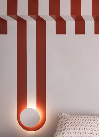 Mur avec décoration en formes géométriques, comprenant des bandes rouges et blanches, avec un éclairage chaud près d'un miroir rond rouge et un oreiller à motifs sur un lit.