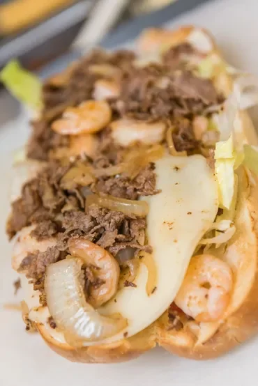 Close-up of a baked seafood and beef sandwich with melted cheese, shrimp, beef, and vegetables in a bun.