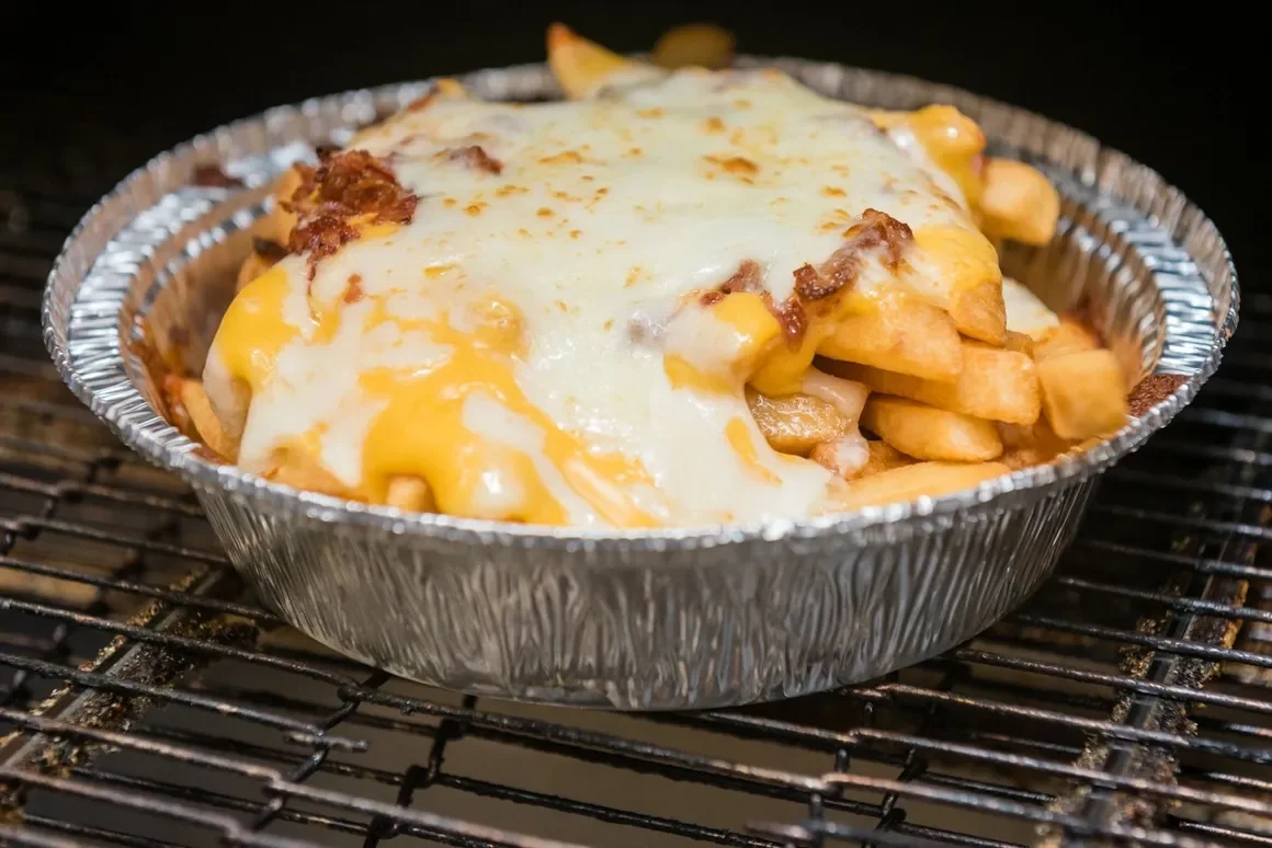 A disposable aluminum container filled with cheesy loaded fries on a grill.