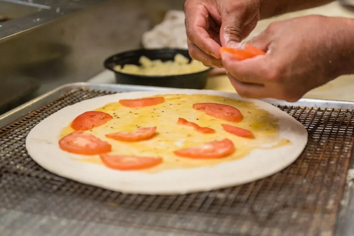 Hands placing sliced tomatoes on a pizza dough topped with cheese.
