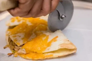 A hand slicing a chicken and cheese burrito with a pizza cutter on a white surface.