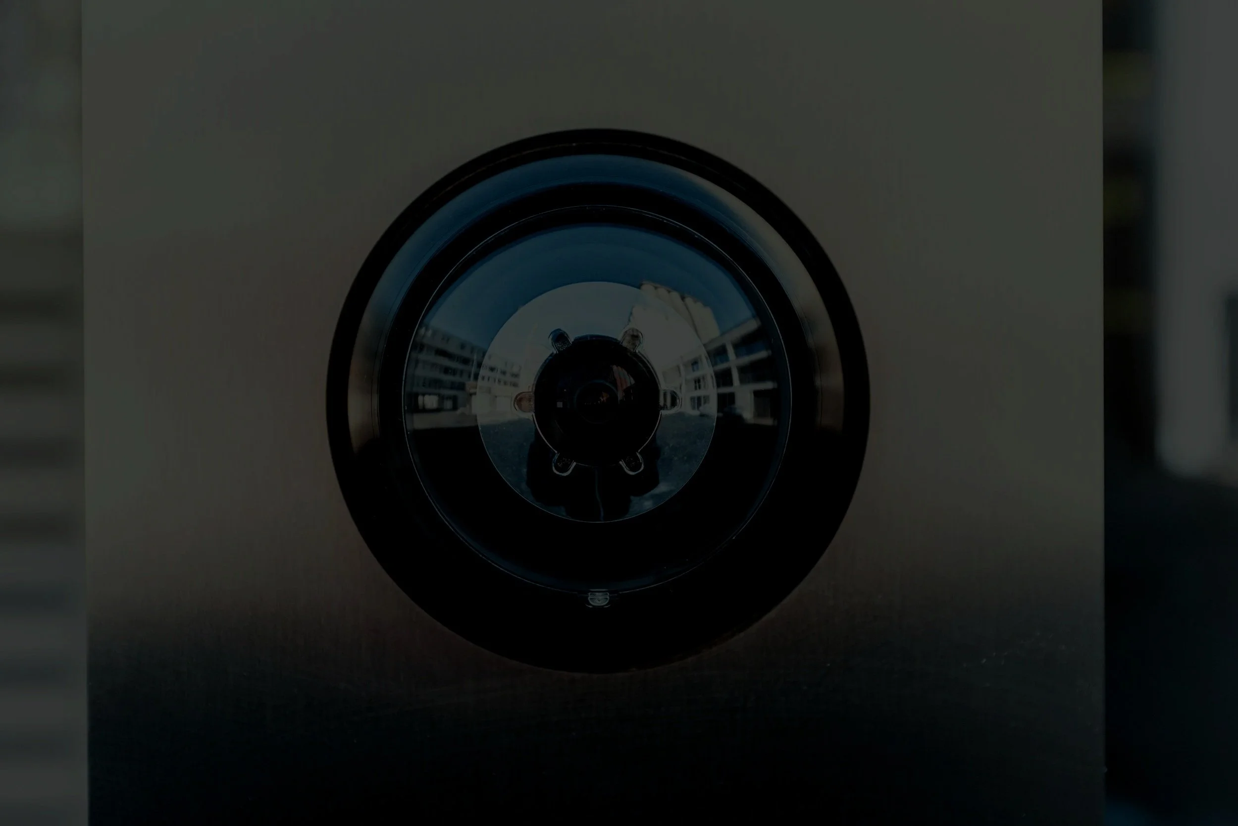 Close-up of a security camera mounted on a wall, with reflections of buildings and a person taking a photo visible on the camera lens.