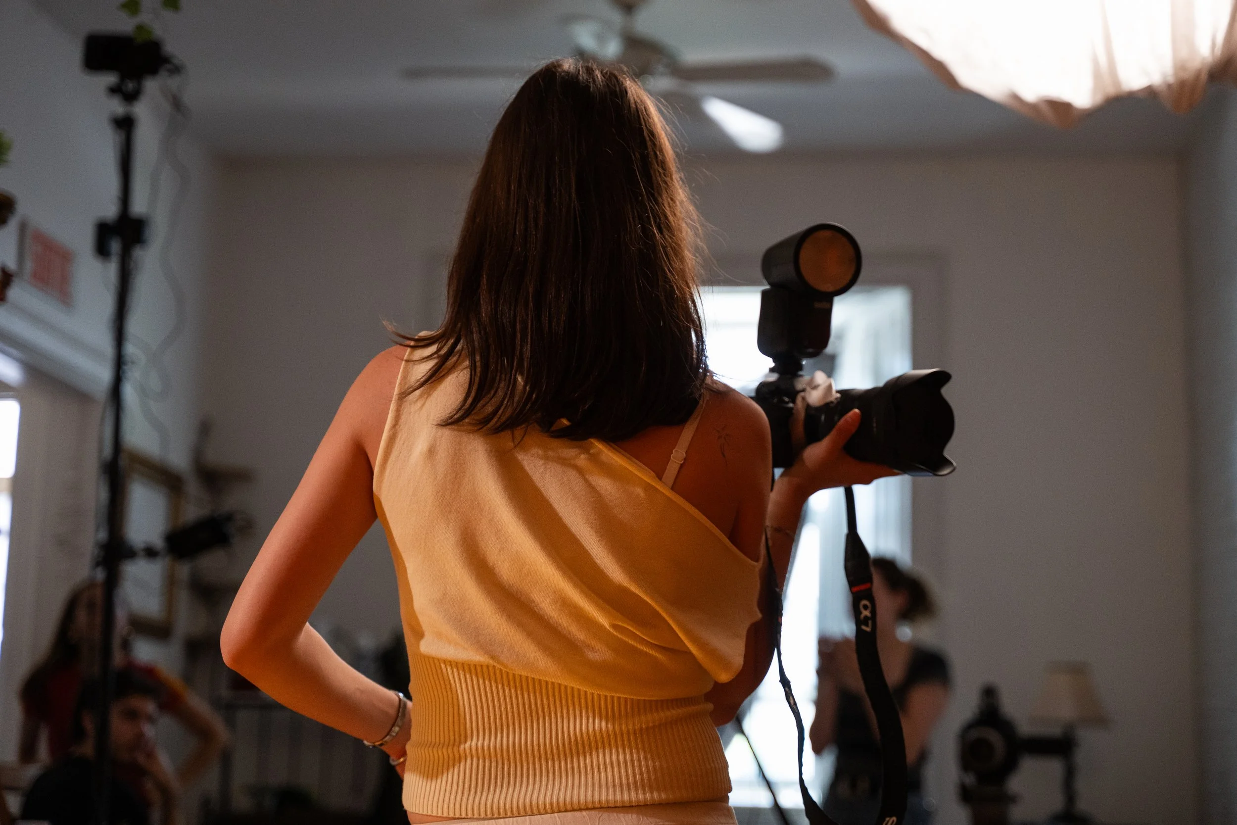 Sienna David, Photographer and Lead Videographer at The Extended Plays. She has shoulder-length brown hair wearing a yellow sleeveless top holding a camera in a room with photography equipment.