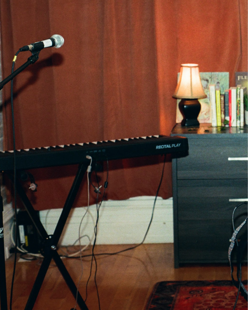 A keyboard on a stand with a microphone on a boom arm in a room with a wooden wall, a lamp, and a small bookshelf.