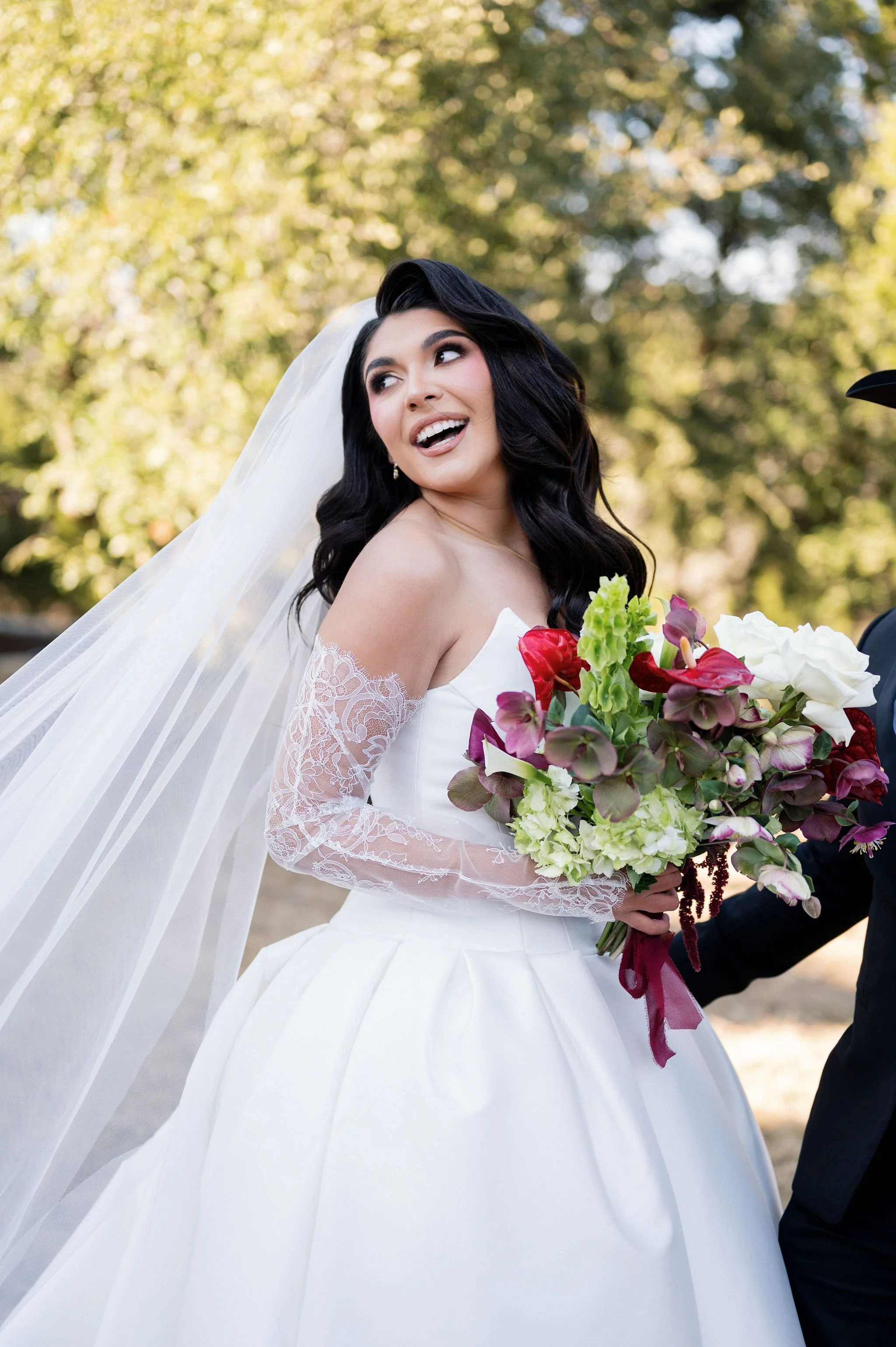 A bride in a white wedding dress with lace sleeves holding a bouquet of flowers, smiling outdoors with blurred trees in the background.
