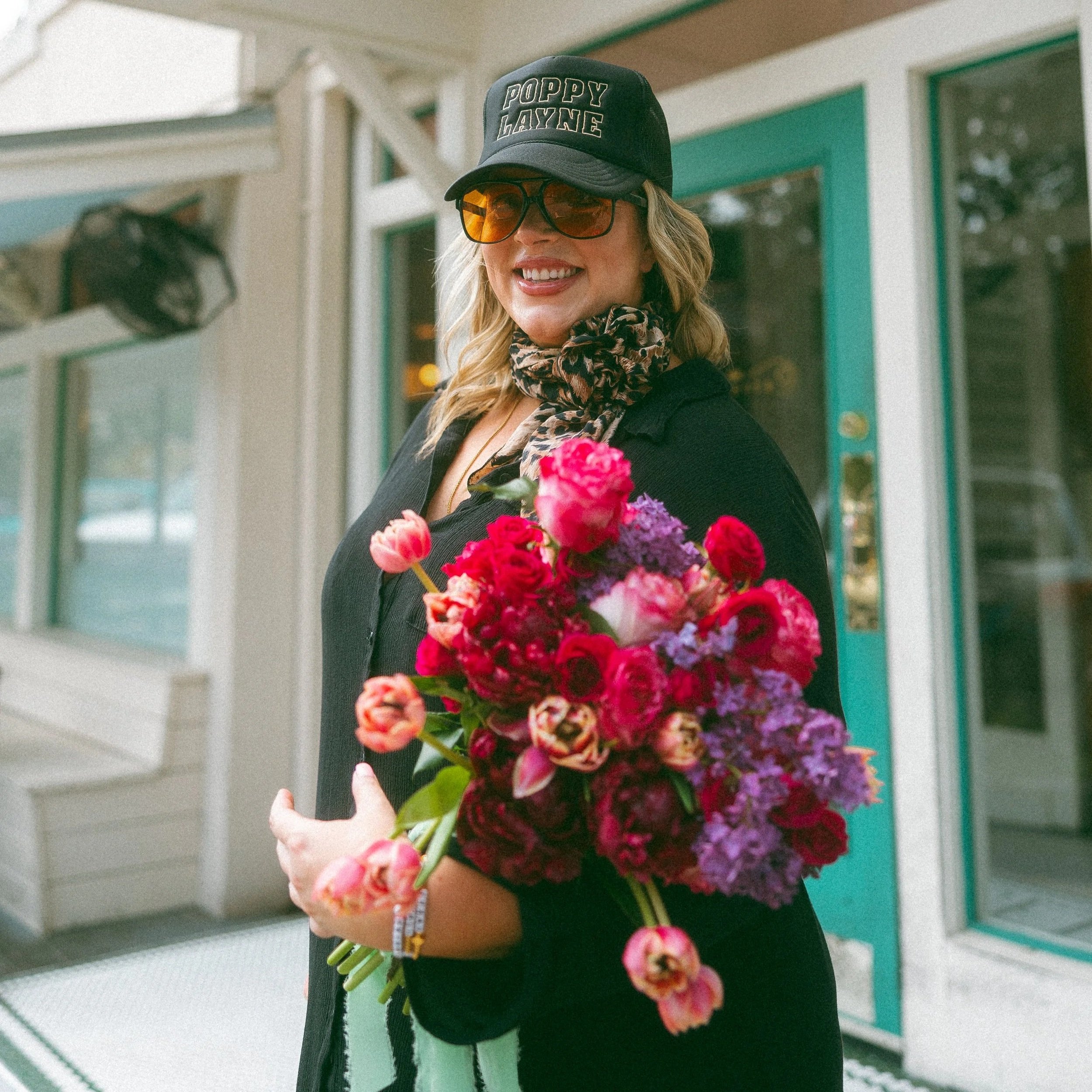 A woman smiling and holding a bouquet of pink, purple, and red flowers outside a building with green window frames.