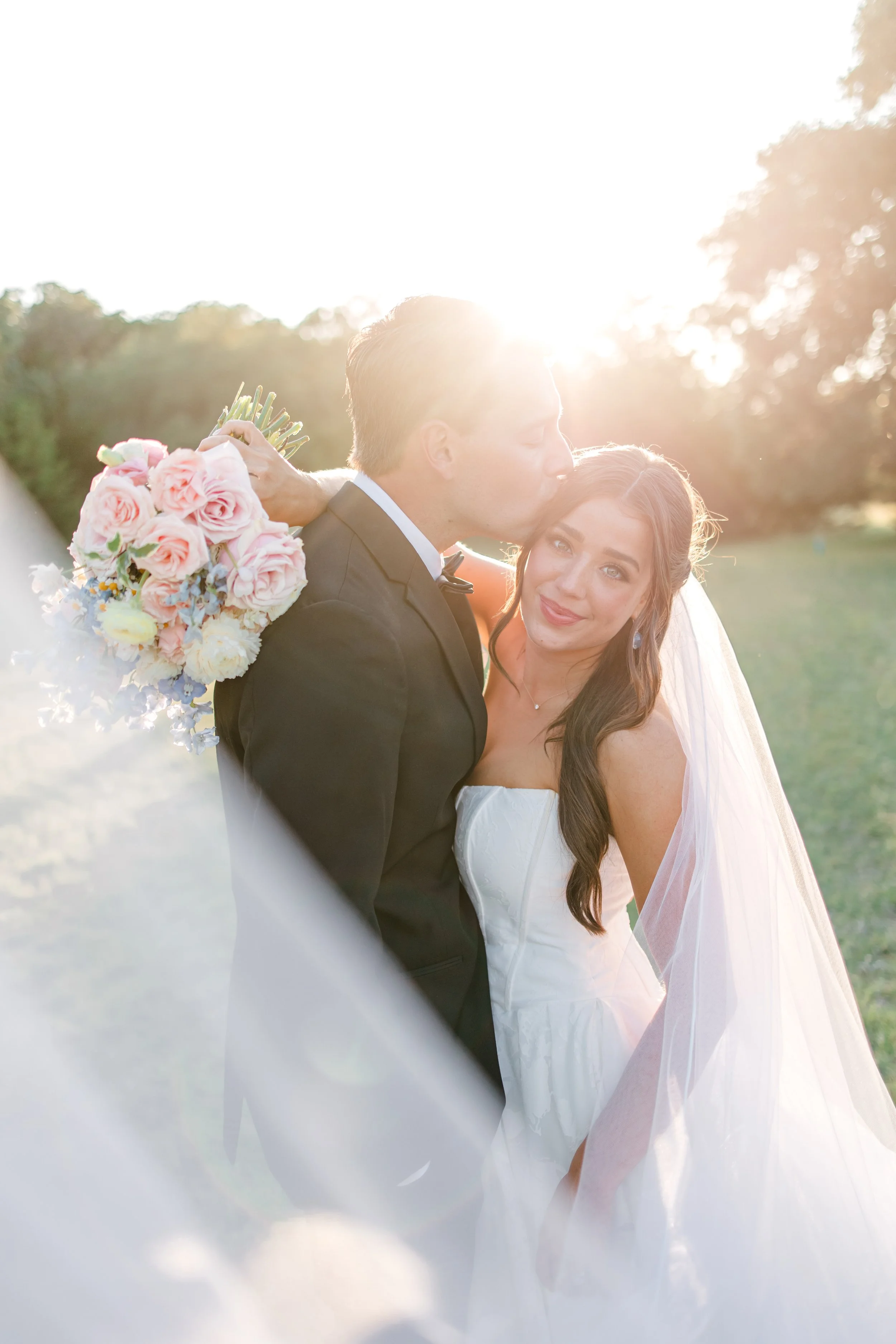 A newlywed couple standing outdoors in sunlight, the groom kissing the bride on the forehead, holding a bouquet of pink and white roses.