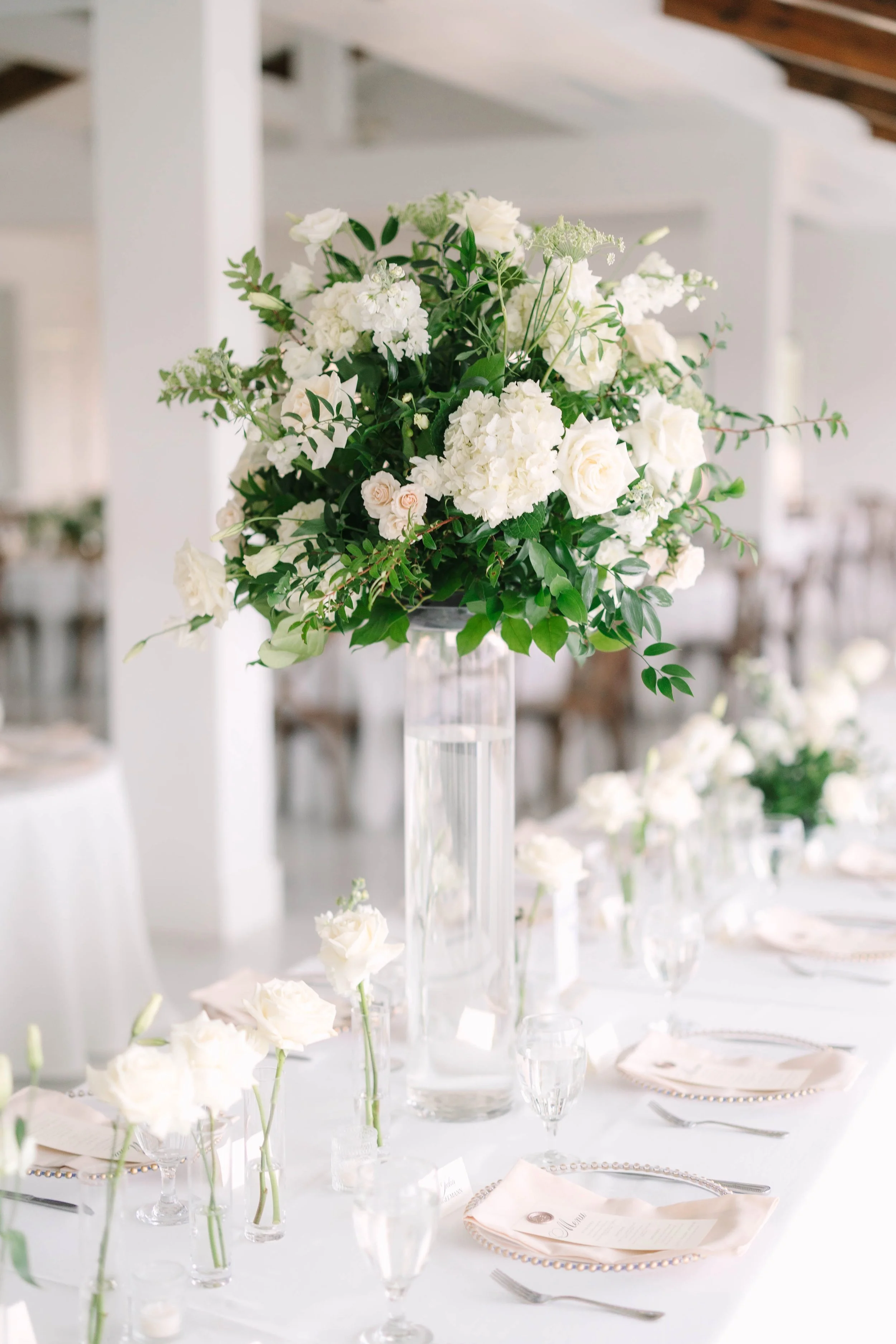 Elegant table setting with a tall floral arrangement of white flowers and greenery in a glass vase, surrounded by small individual flower arrangements and place settings with white napkins and glassware.