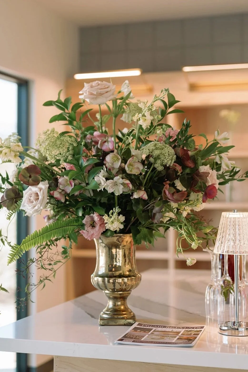 A large flower arrangement with pink, white, and green flowers in a gold vase on a white countertop, with a lamp and a magazine nearby in a modern room.