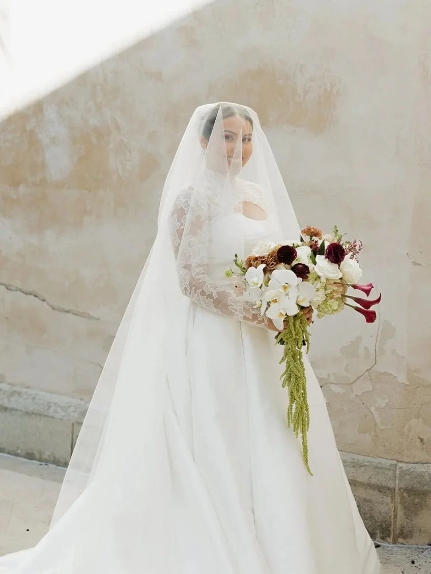 Bride in a white wedding dress and veil holding a bouquet of white and dark red flowers, standing against a beige wall.