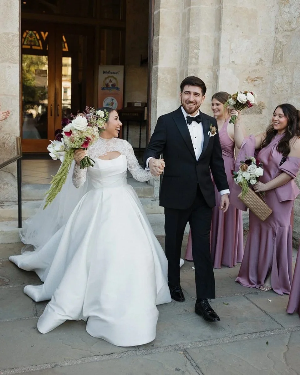 A newlywed couple holding hands and smiling outside a building, with three bridesmaids in lavender dresses holding bouquets behind them.